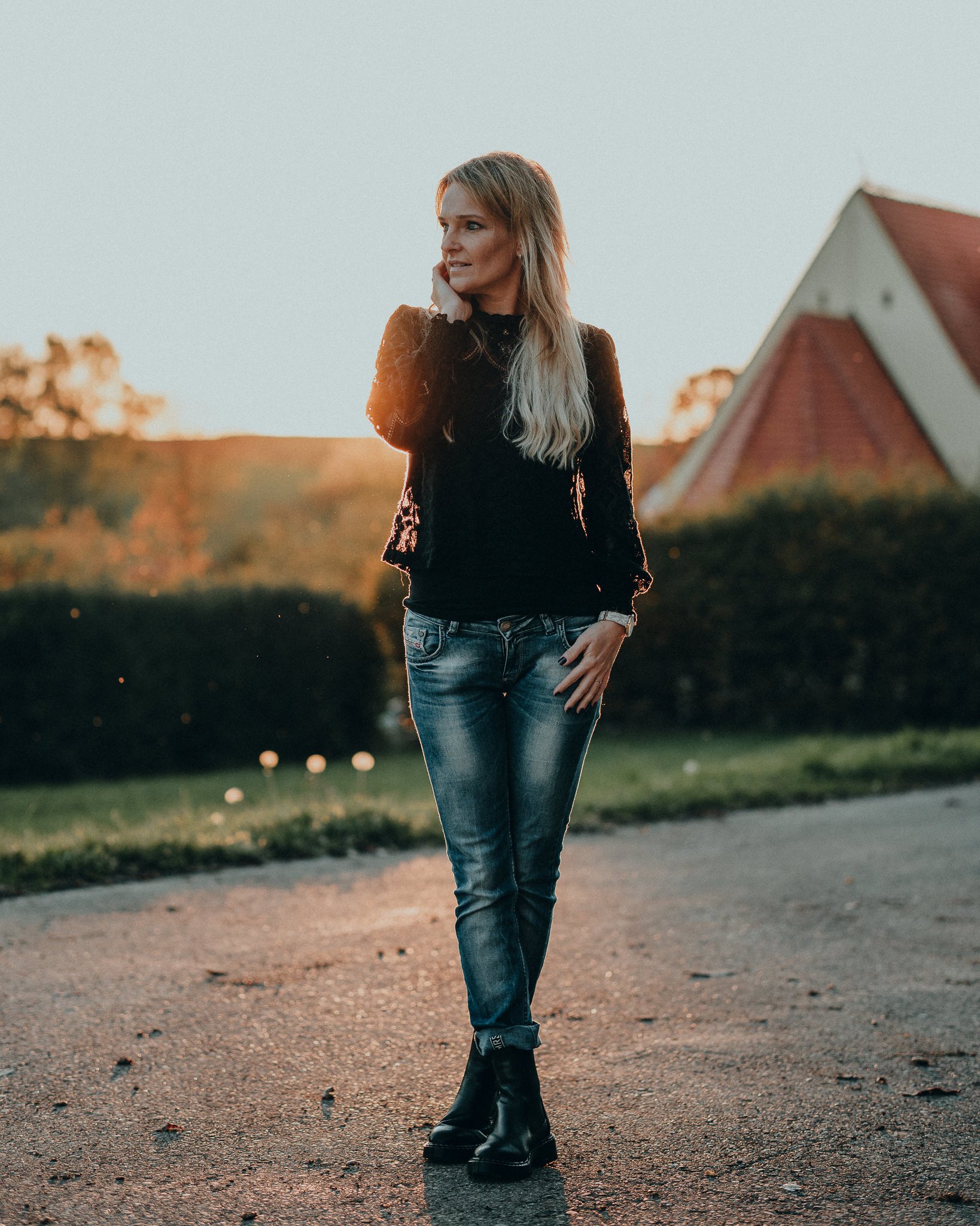 Junge Frau steht auf einer Straße bei Sonnenuntergang, trägt eine schwarze Bluse, Jeans und schwarze Stiefel, mit einer Hand im Haar, im Hintergrund ist ein Haus mit rotem Dach.