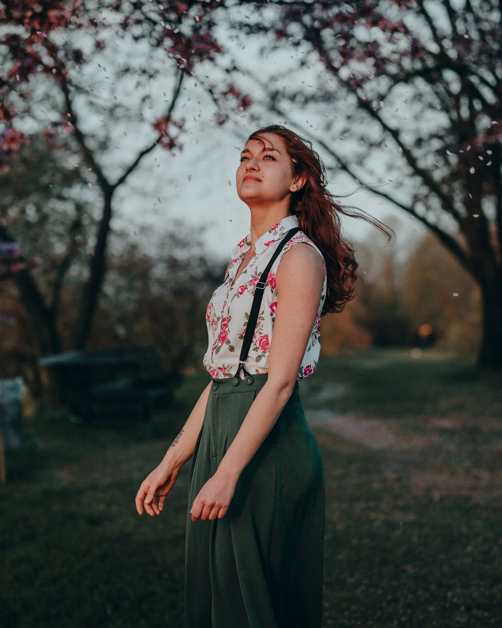 Eine junge Frau steht im Freien unter blühenden Bäumen, wirkt entspannt und selbstsicher, trägt einen weißen Top mit Blumenmuster, grüne hohe Hosen und hat langes braunes Haar, das im Wind weht.