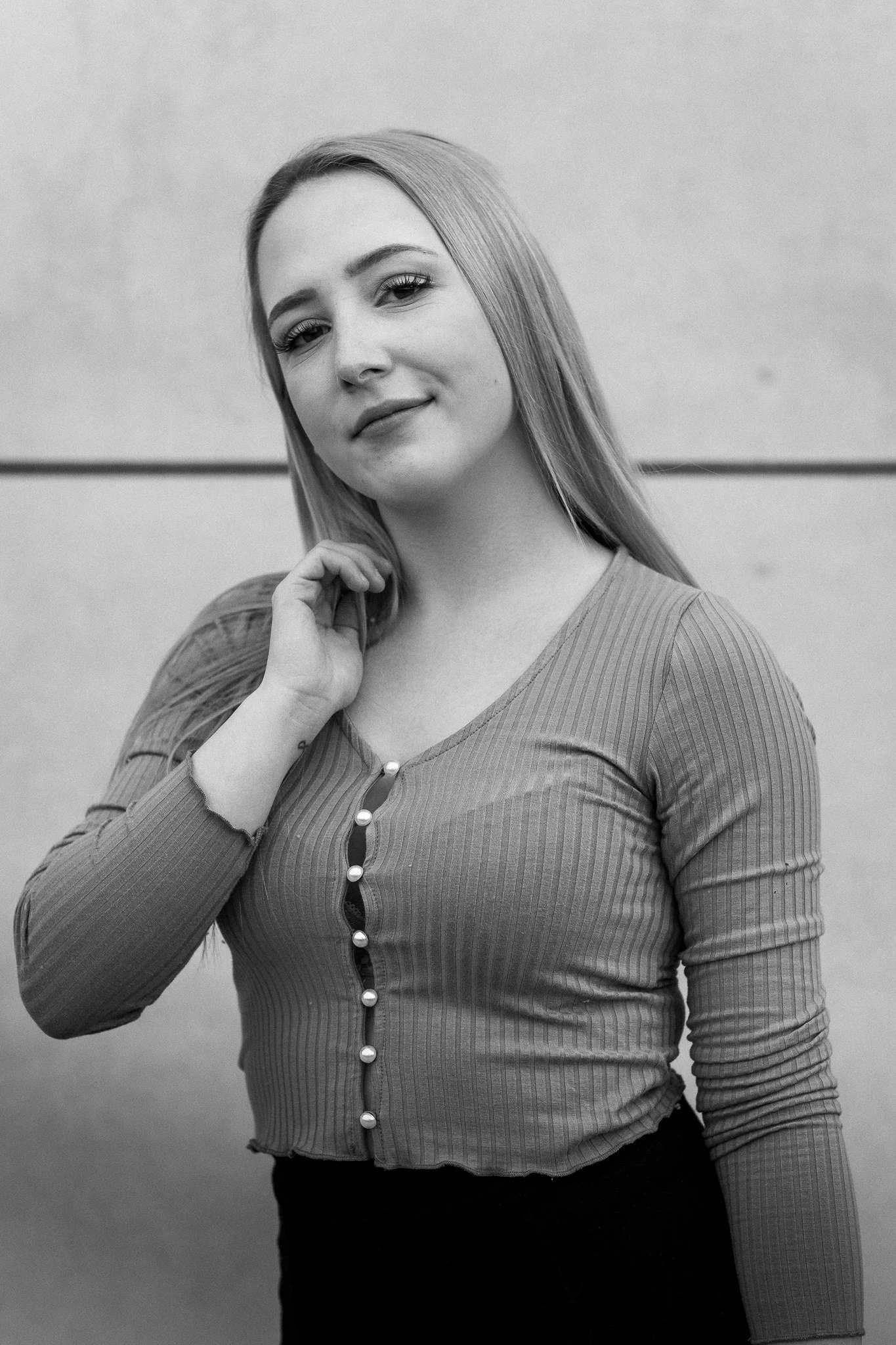 Schwarz-weiß-Fotografie einer jungen Frau mit langem Haar, die nach rechts schaut, die Hand an ihrem Hals und leicht lächelt, vor einer glatten Wand.