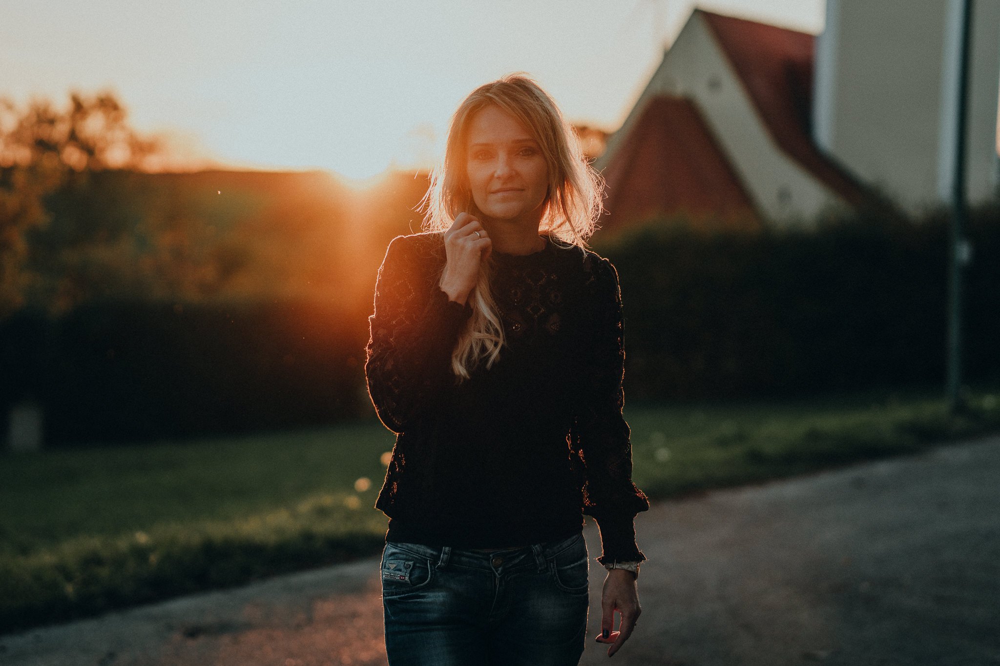 Junge Frau steht im Abendlicht vor Häusern, trägt eine schwarze Spitzebluse und Jeans, beim Sonnenuntergang.