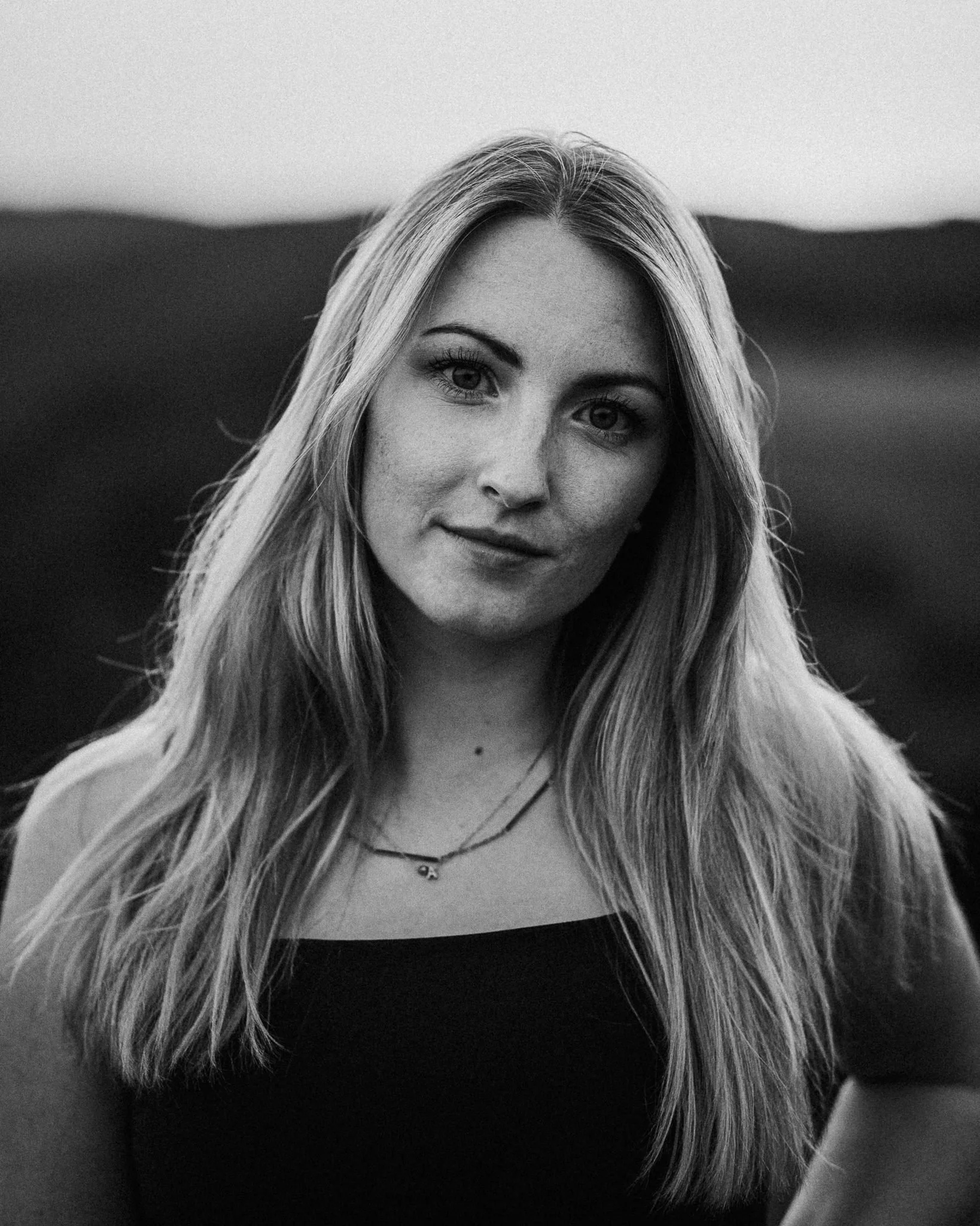 Schwarz-weiß Porträt einer jungen Frau mit langen, glatten Haaren, hellem Hauttyp und einem dezenten Lächeln, im Freien mit unscharfem Hintergrund.