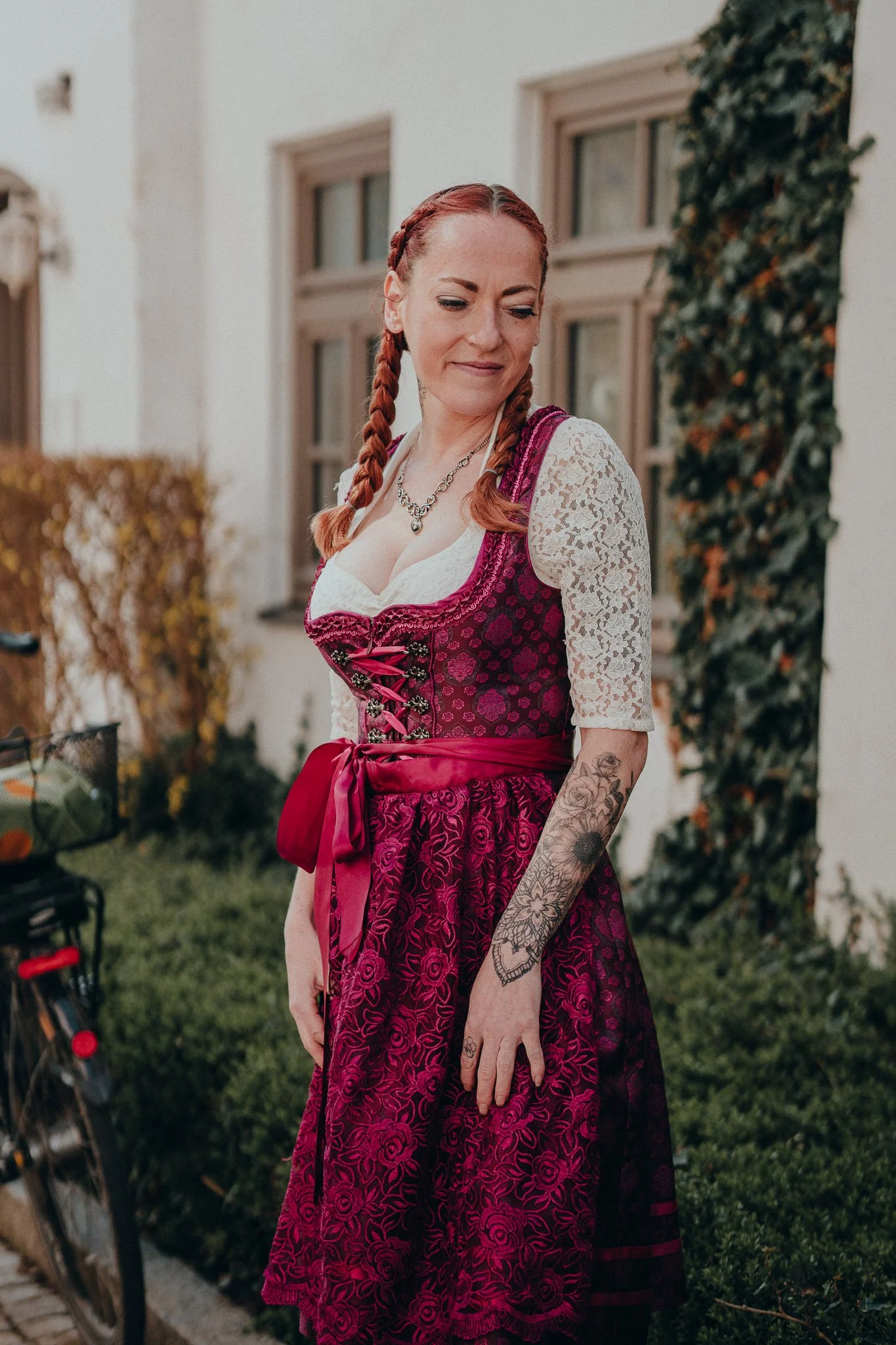 Frau in traditionellem Dirndl mit roten Zöpfen, Tattoos an der Hand, steht vor einem Haus mit Efeuranken, blickt seitlich.
