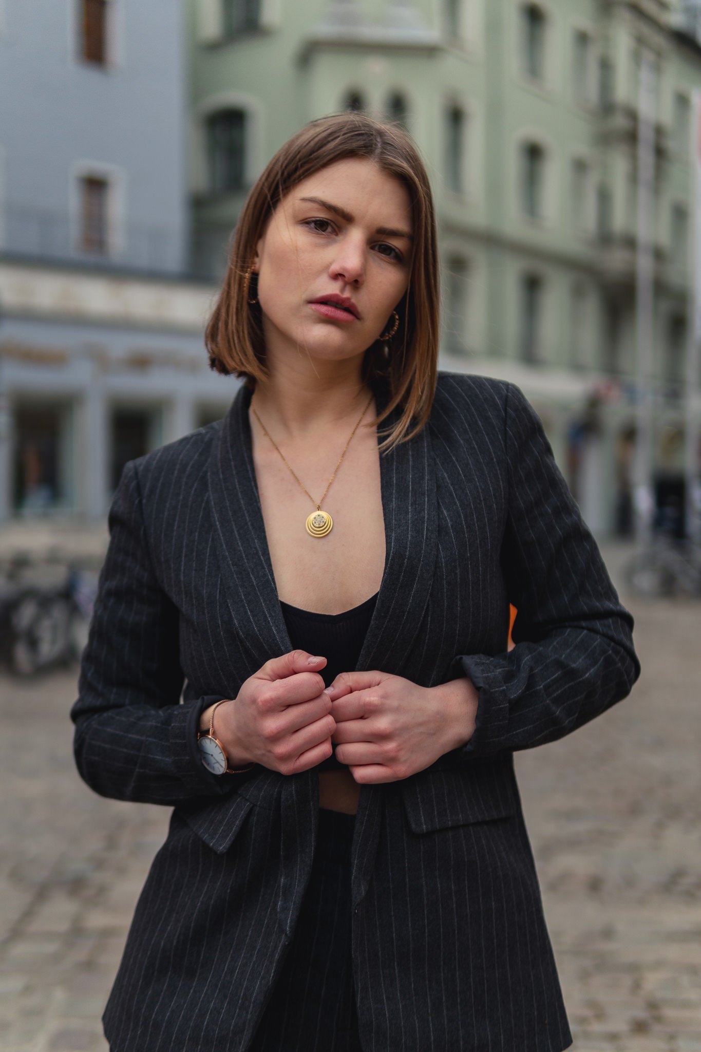 Junge Frau in dunklem Anzug steht vor urbaner Kulisse, trägt Halskette und Uhr, schaut ernst in Kamera.