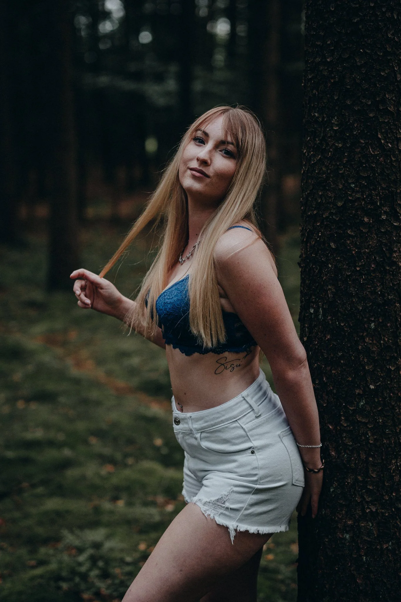 Jugendliche Frau mit langem blondem Haar lehnt an einem Baum im Wald, trägt blaue Spitze-BH und weiße Shorts, schaut in die Kamera.