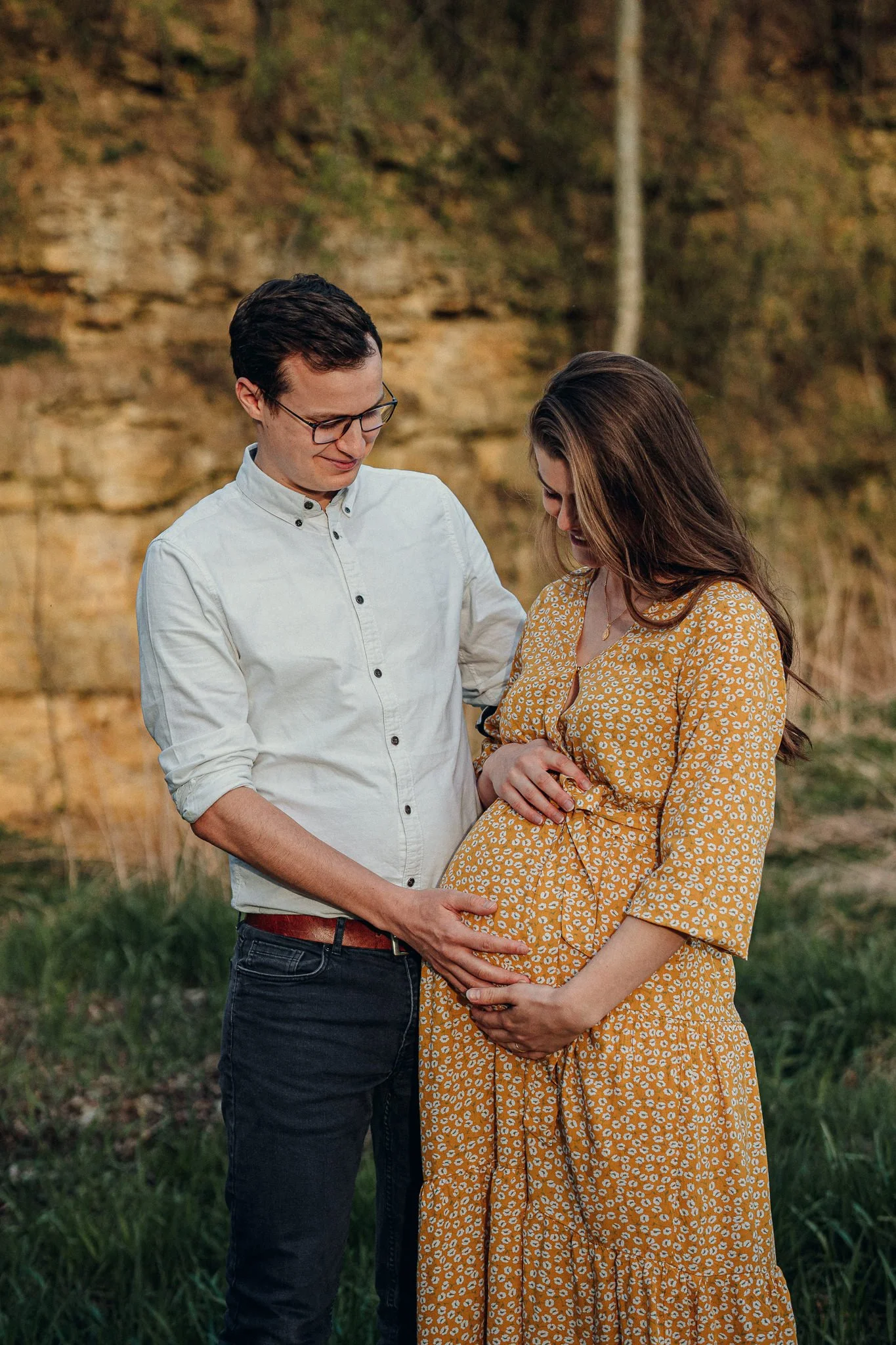 Ein Paar hält die Hand auf den Bauch der schwangeren Frau, die lächelnd nach unten schaut, im Freien bei Sonnenuntergang.