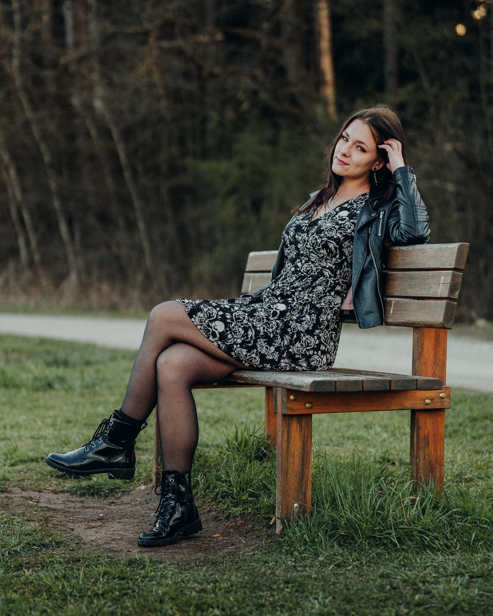 Junge Frau sitzt auf einer Parkbank im Grünen, trägt ein schwarzes Kleid mit weißen Blumenmustern, schwarze Stiefel und eine schwarze Lederjacke, hat braune Haare und sieht in die Kamera.