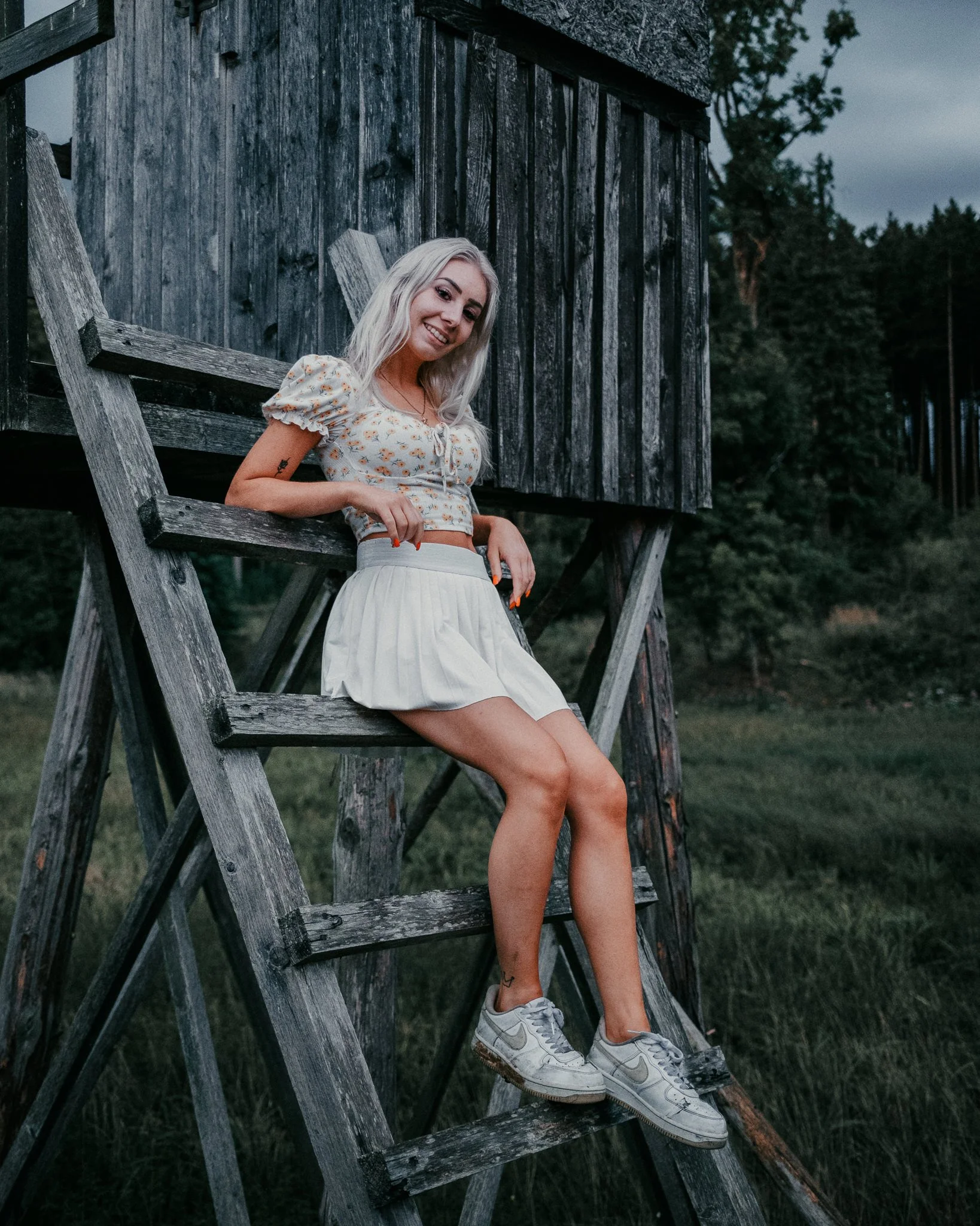 Junge Frau sitzt auf einer Leiter neben einer Holzhütte im Freien, umgeben von Bäumen, bei bewölktem Himmel.