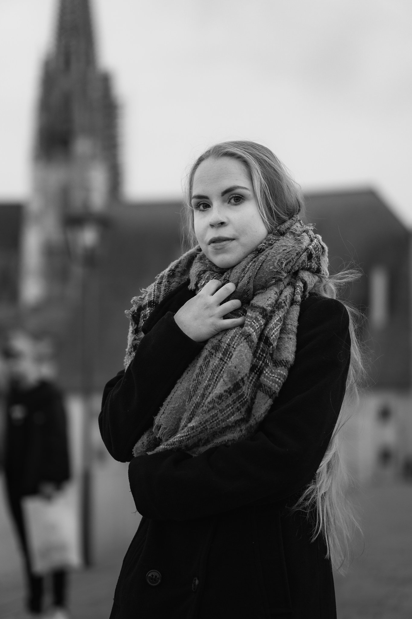 Schwarz-Weiß-Foto einer jungen Frau mit langen, lockigen Haaren, die einen karierten Schal trägt und vor einem unscharfen Hintergrund steht.