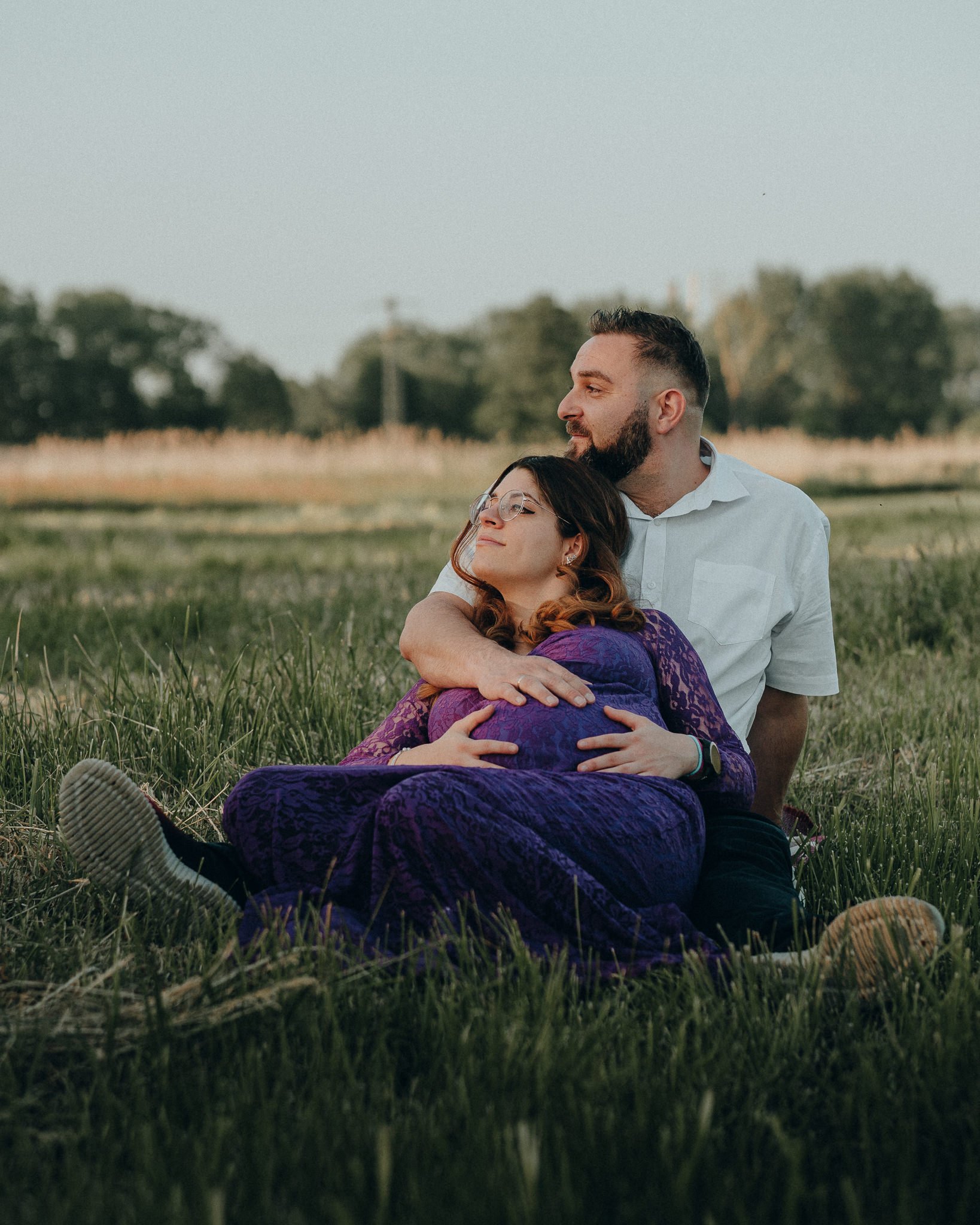 Ein Paar sitzt im Grünen, umarmt sich liebevoll, Frau trägt eine lila Kleide und trägt eine Brille, Mann trägt ein weißes Hemd. Es ist draußen im Freien, vermutlich am Abend.