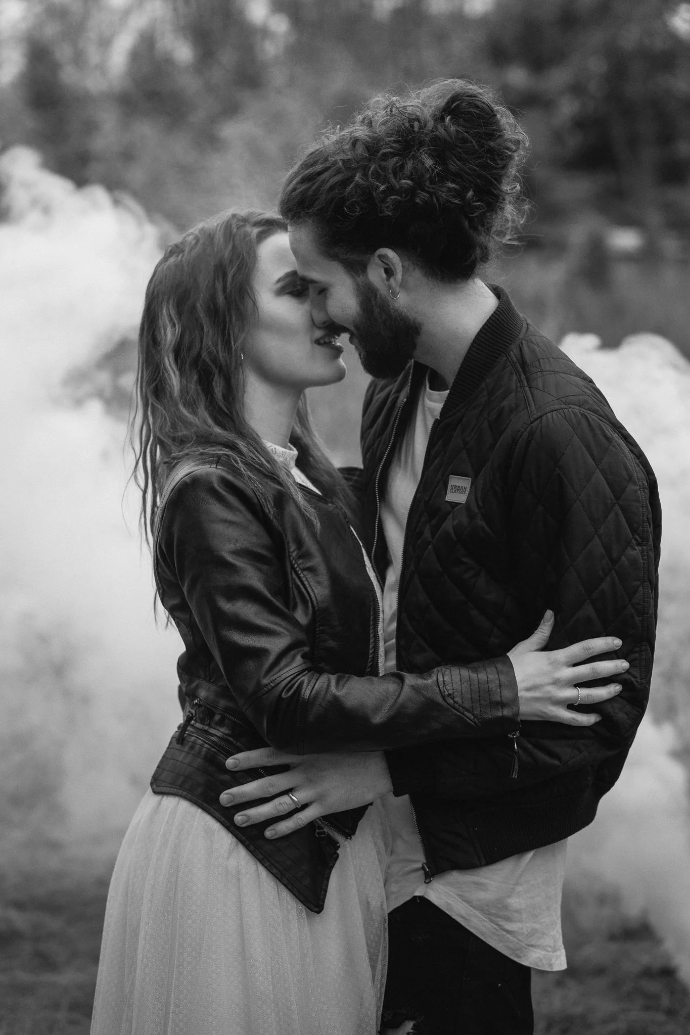 Ein verliebtes Paar steht sich in einer natürlichen Umgebung nahe einem Wasser, ihre Stirn und Nasen berühren sich in einem Kuss, beide lachen und tragen dunkle Jacken.