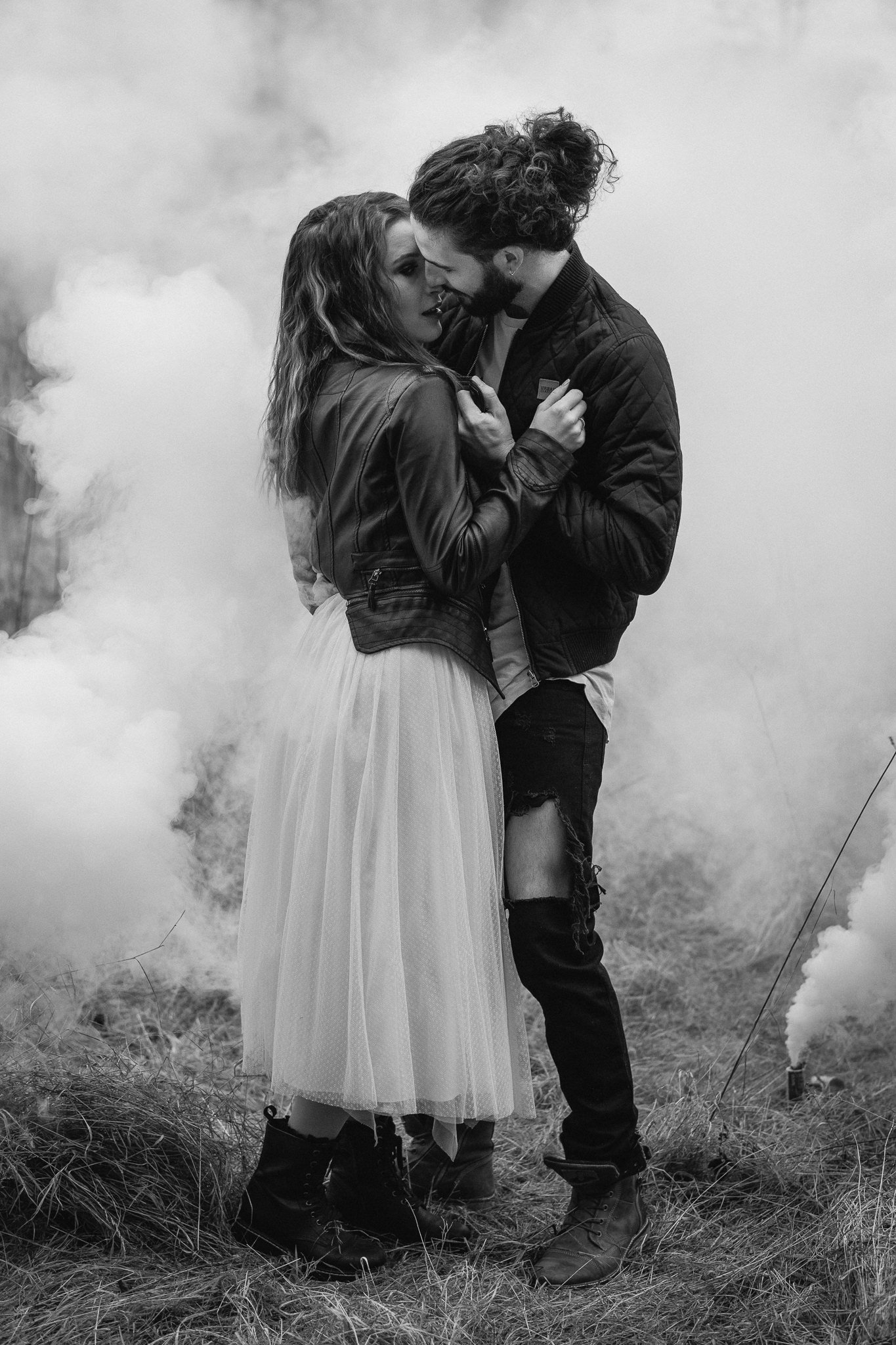 Ein Paar steht sich nahe beieinander, umgeben von Rauchschwaden, in einer offenen, grasbewachsenen Fläche.