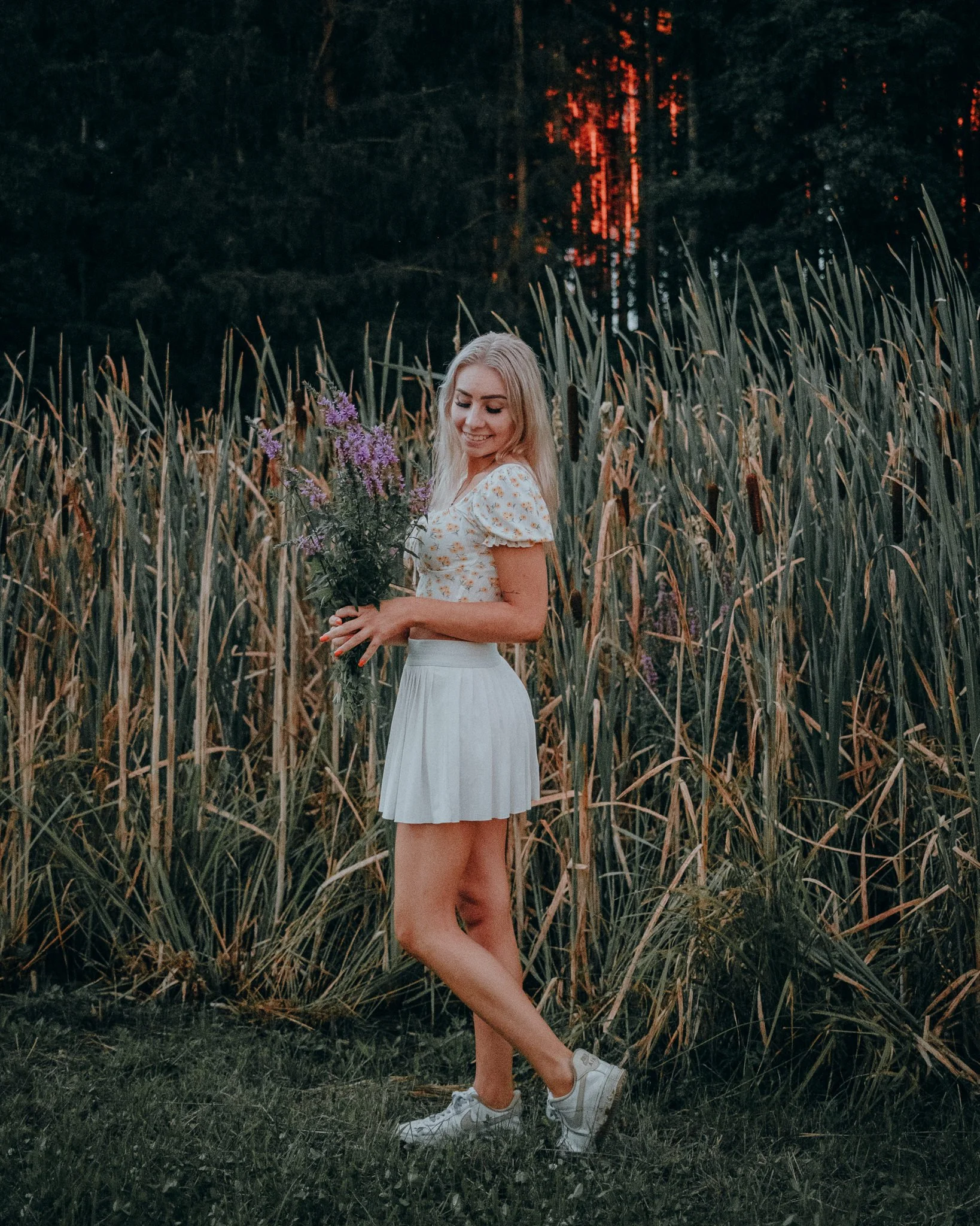 Eine junge Frau hält Blumen in einem Feld bei Sonnenuntergang, trägt ein weißes Kleid und Sneakers.