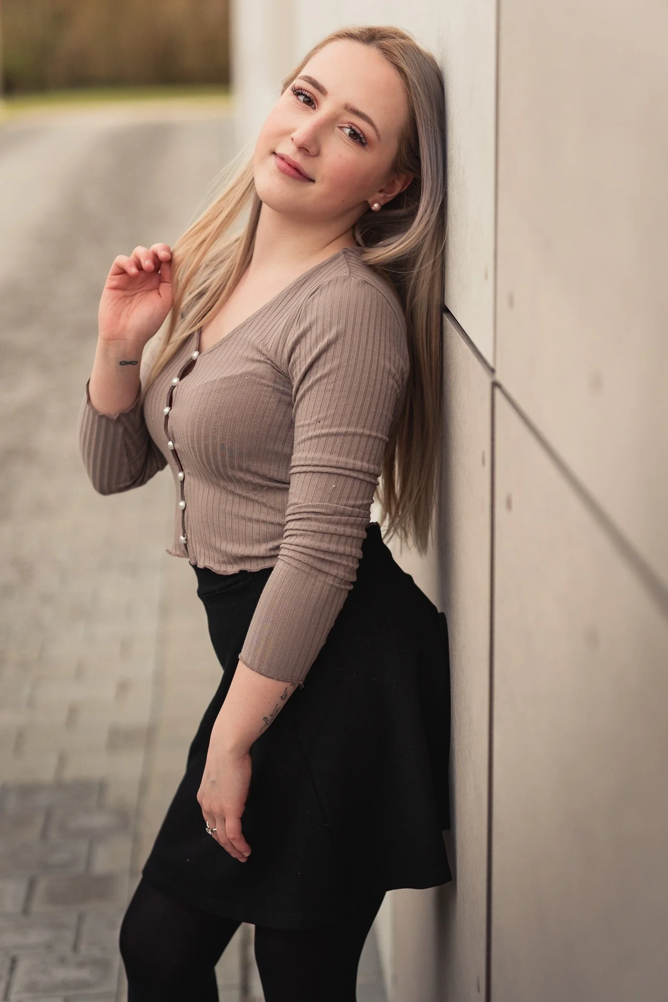 Junge Frau lehnt an einer Wand im Freien, trägt eine braune, begrenzte Oberteil und eine schwarze Rock, hat lange blonde Haare und lächelt leicht.