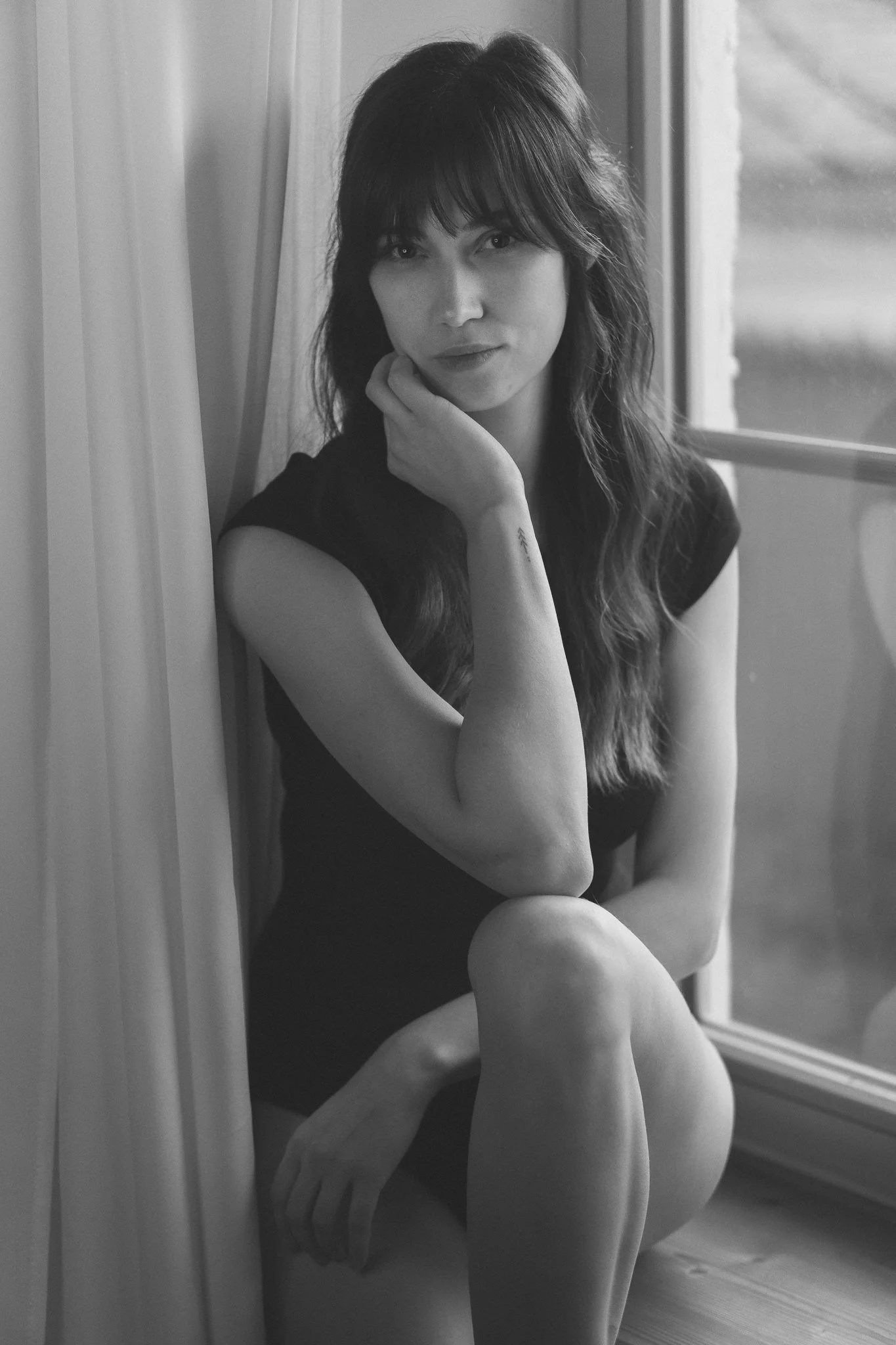 Schwarz-weiß-Foto einer jungen Frau mit dunklen, gewellten Haaren, die neben einem Fenster sitzt, die Hand am Kinn. Sie trägt ein kurzes, dunkles Kleid.