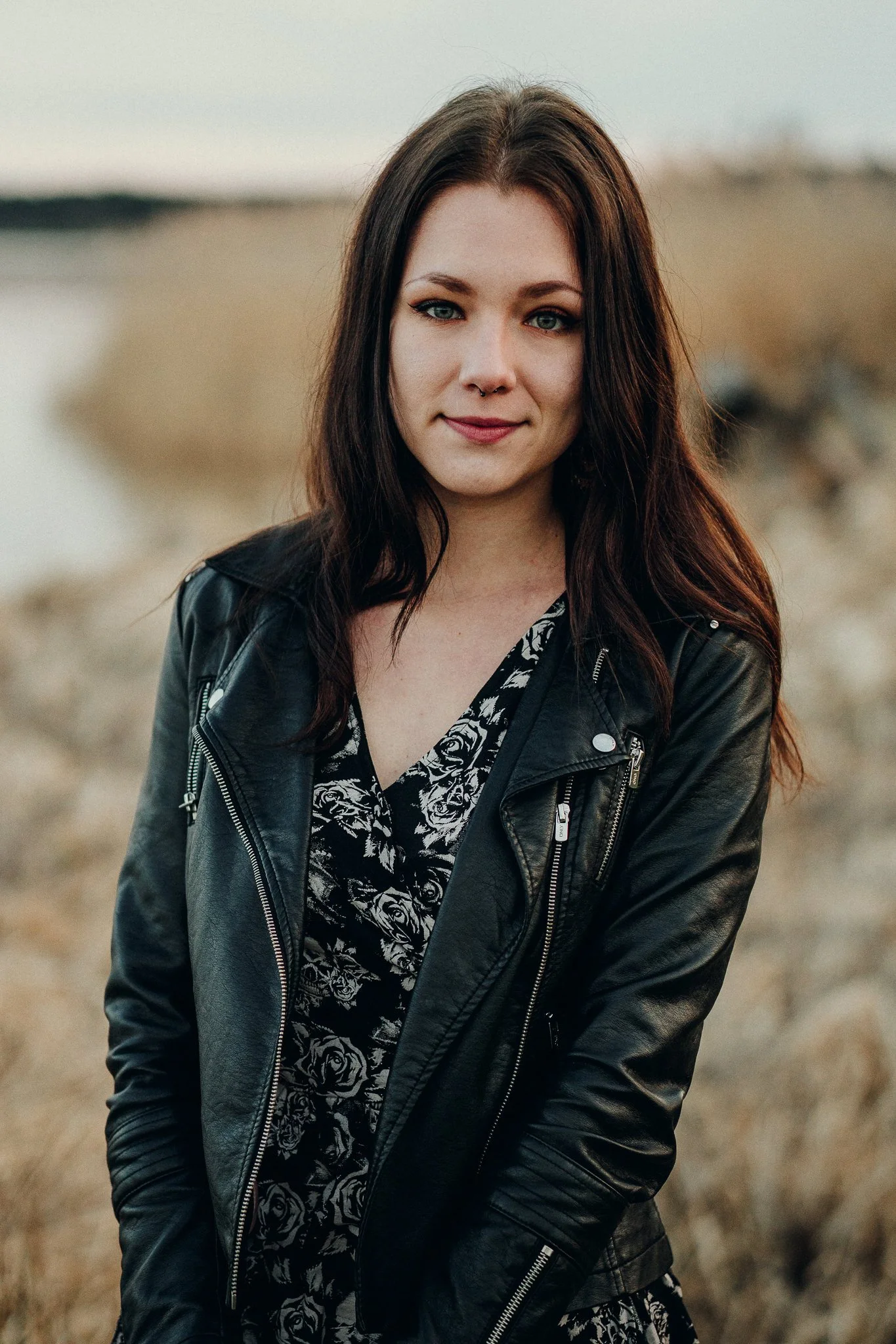 Ein junges Mädchen mit braunen, welligen Haaren steht in einer natürlichen Umgebung mit einer Wasserfläche im Hintergrund. Sie trägt eine schwarze Lederjacke und ein schwarzes Kleid mit weißen Rosenmuster.