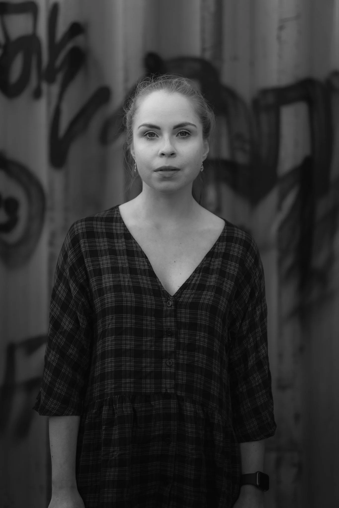 Schwarz-weiß Porträt eines jungen Mädchens mit gedrückten Haaren, das einen karierten Kleider trägt, vor einer Holzwand mit künstlerischen Graffiti. Sie schaut direkt in die Kamera.