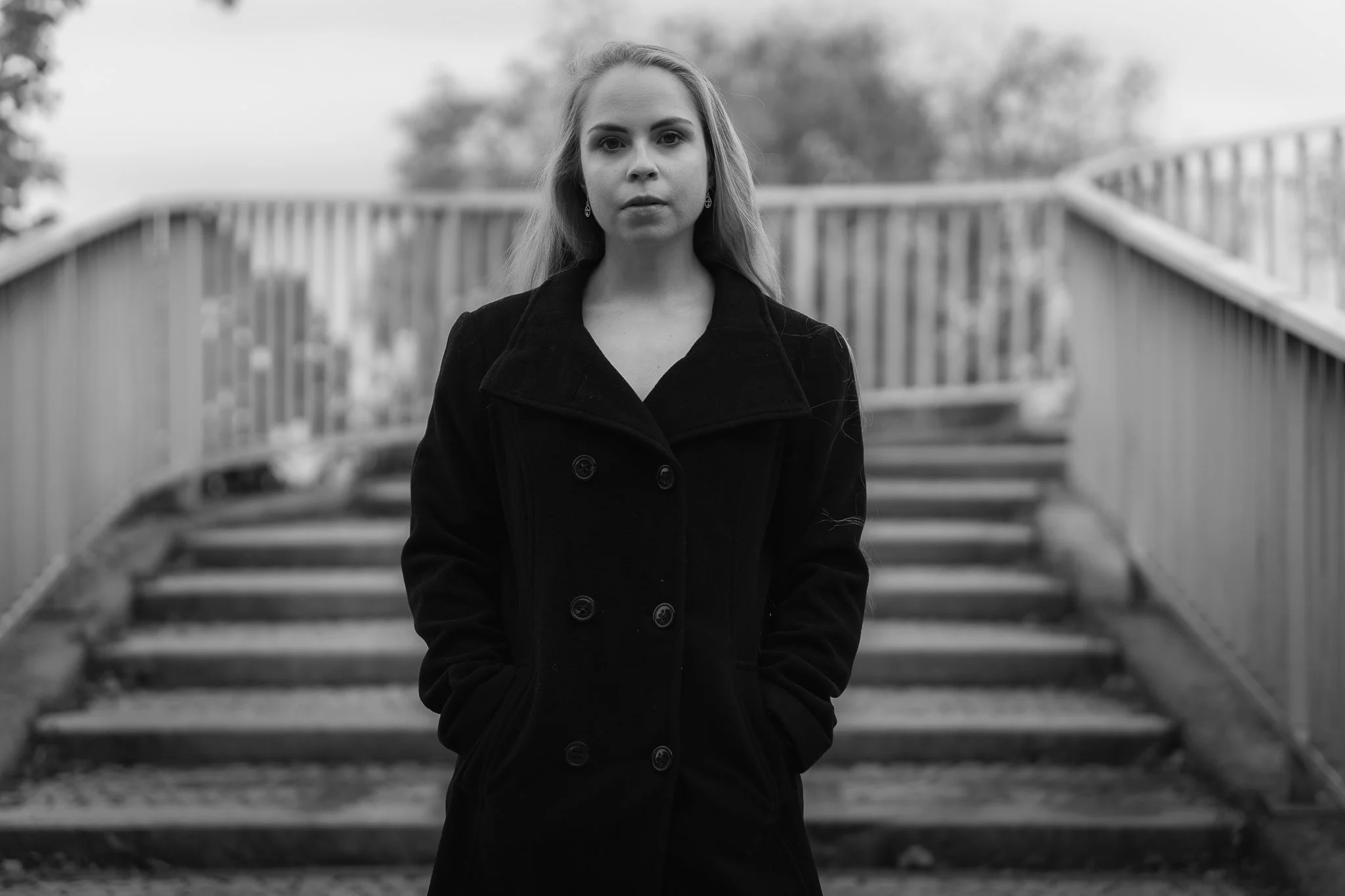 Junge Frau mit langen Haaren trägt einen schwarzen Mantel steht auf einer Brücke, im Hintergrund Bäume, in Schwarz-Weiß fotografiert.