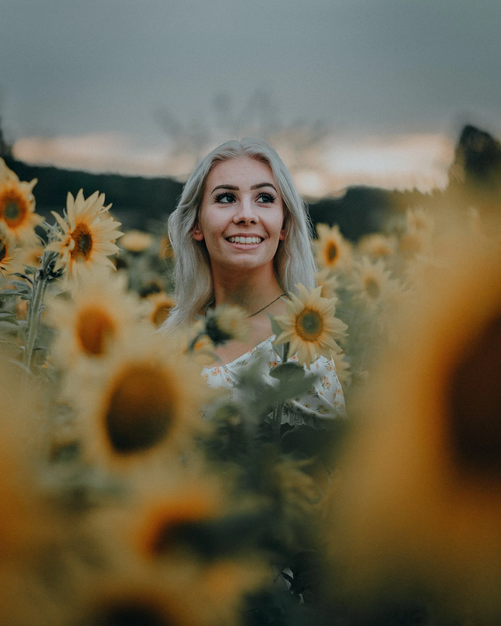 Ein lächelnder junger Mann mit langen, blonden Haaren, umgeben von Sonnenblumen im Freien bei Sonnenuntergang.