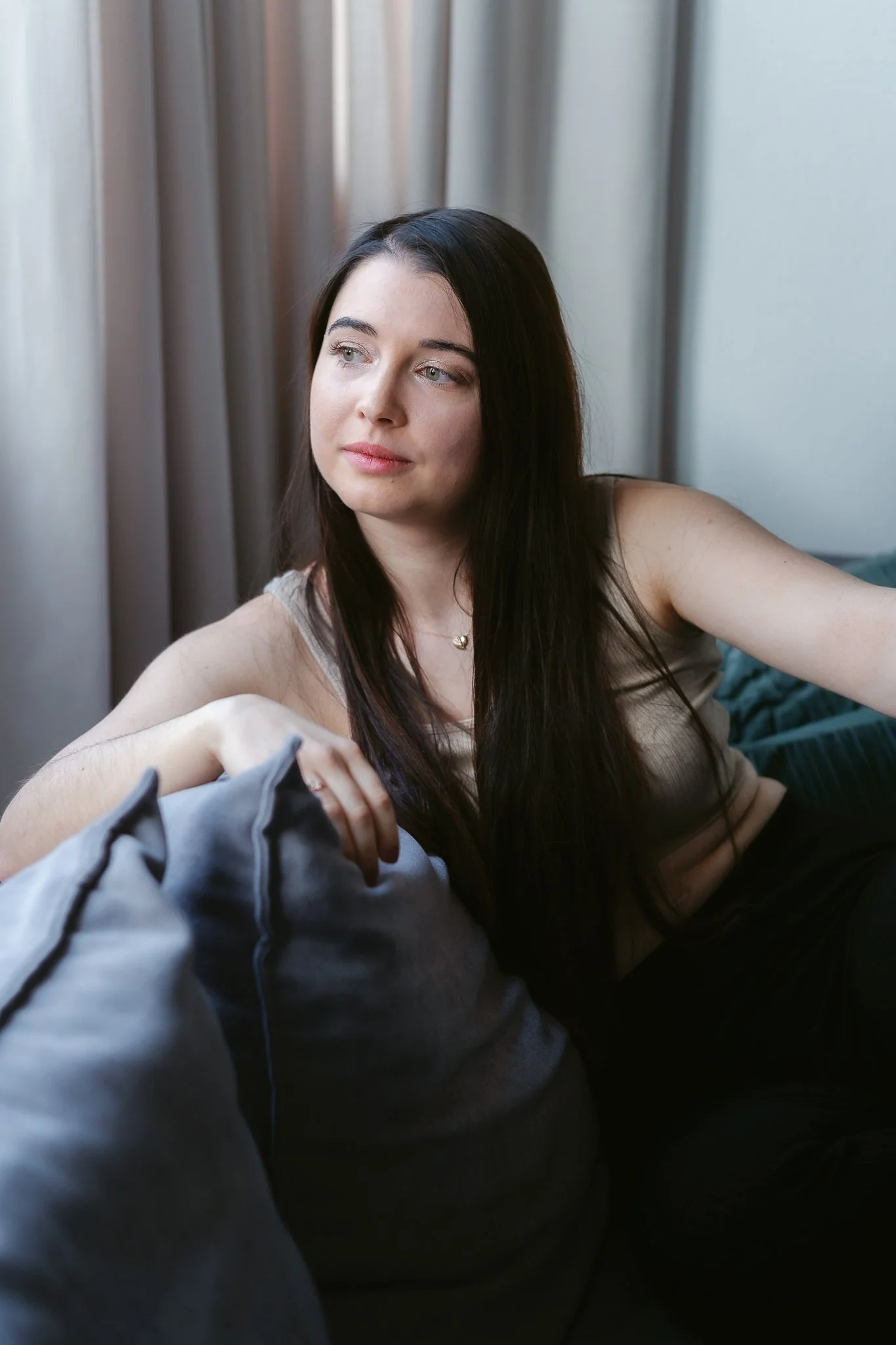 Eine junge Frau mit langen dunklen Haaren sitzt auf einem Sofa, blickt nach links und wirkt nachdenklich.