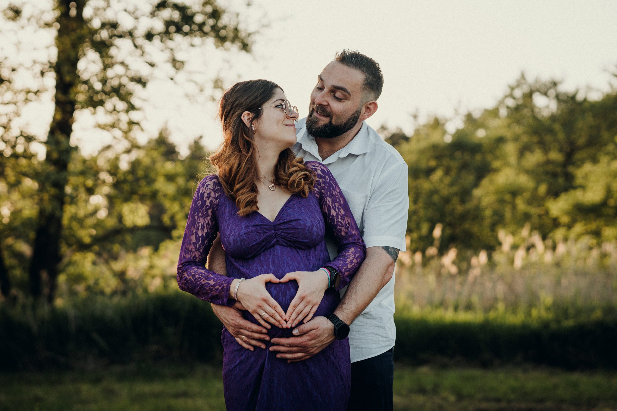 Ein Paar, ein werdendes Elternpaar, steht im Freien in der Abenddämmerung, umarmt sich und macht ein Herz mit den Händen über dem Babybauch, während sie sich liebevoll anschauen.
