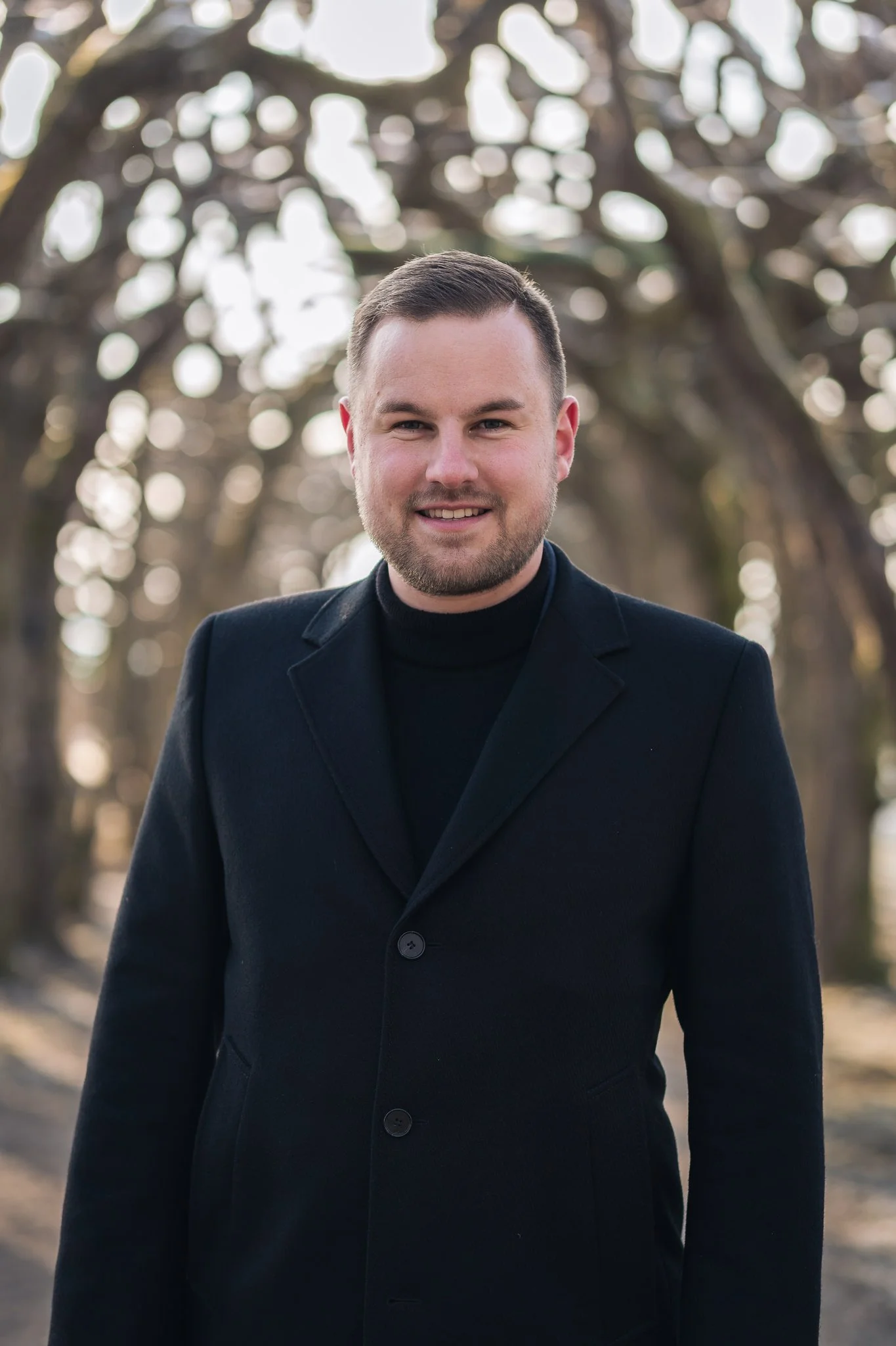 Ein Mann im schwarzen Mantel steht vor einer Baumkulisse im Freien.