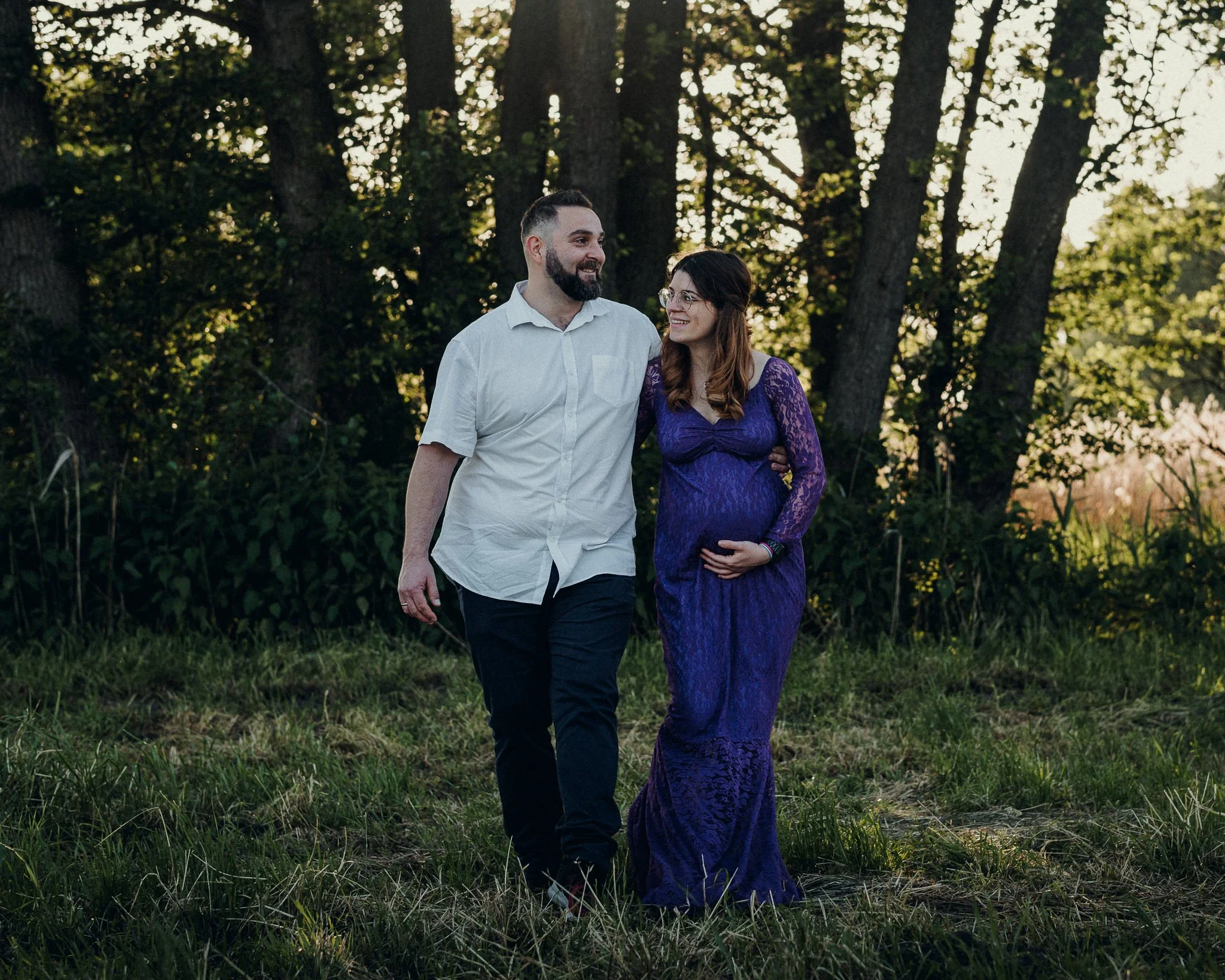 Ein Paar, ein schwangere Frau in einem langen lila Kleid und ein Mann in einem weißen Hemd, geht gemeinsam im Grünen spazieren, umgeben von Bäumen bei Sonnenuntergang.