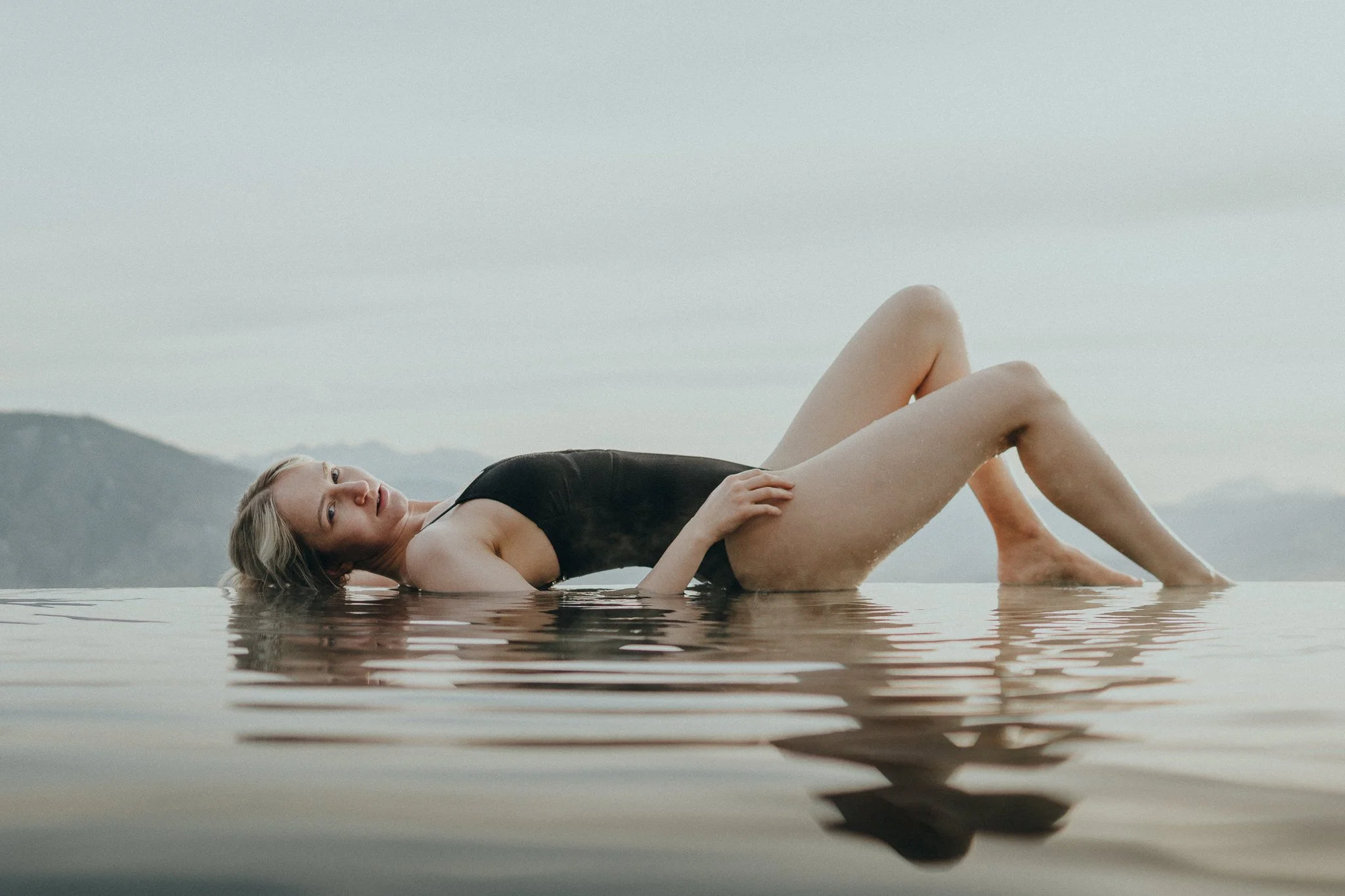 Frau liegt in einem Wasserpool, trägt schwarzen Badeanzug, mit Bergen im Hintergrund bei bewölktem Himmel.