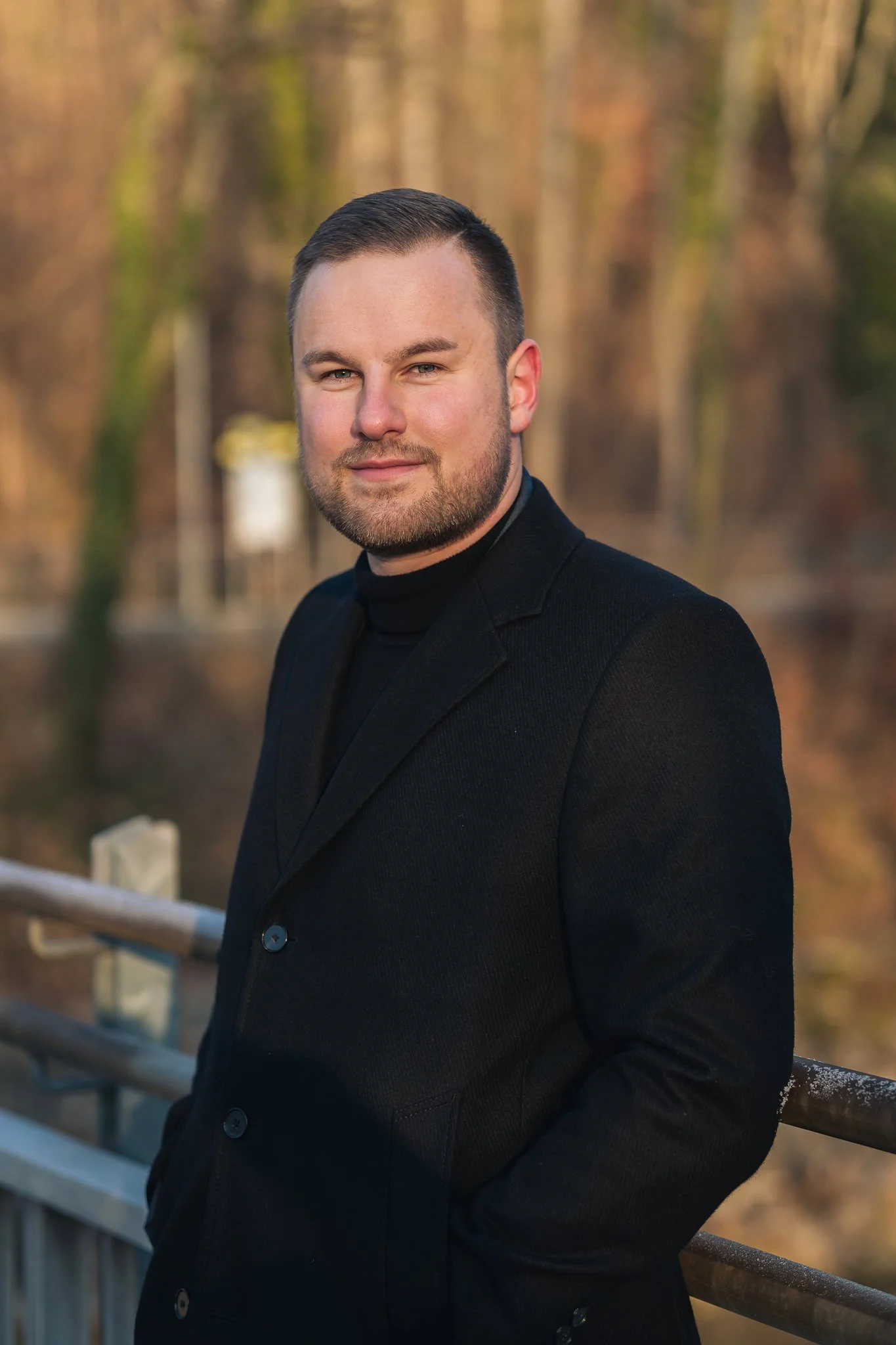 Ein Mann trägt einen schwarzen Mantel und steht an einem Geländer im Freien vor einem herbstlichen Wald. Er lächelt leicht und blickt in die Kamera.