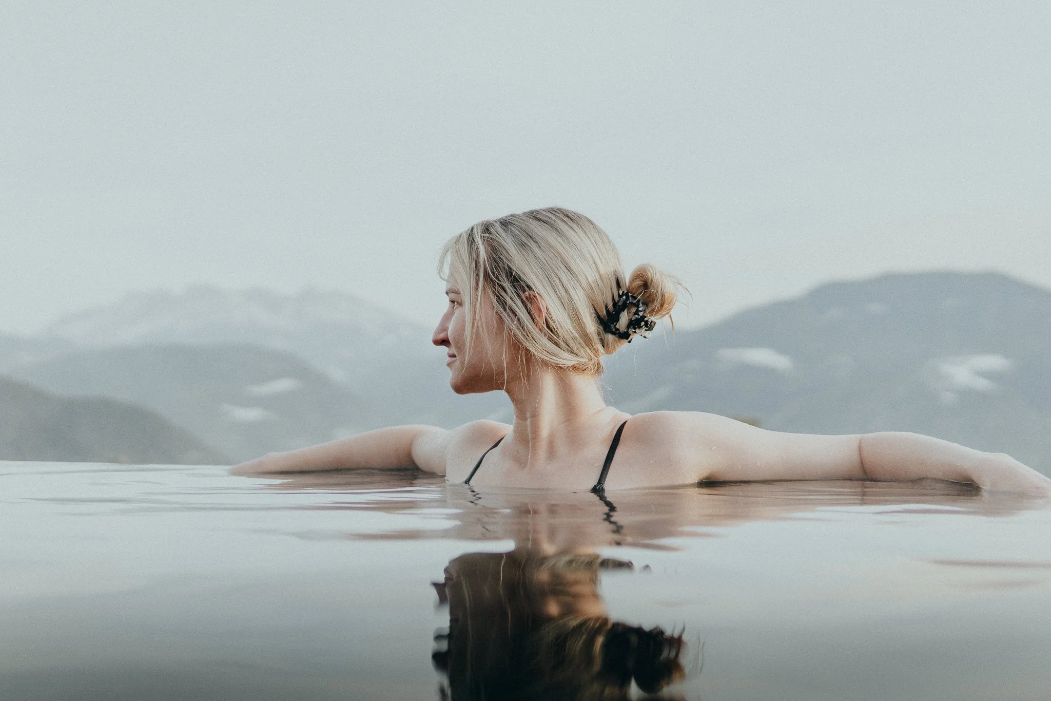 Eine blonde Frau entspannt im Infinity-Pool mit Bergkulisse im Hintergrund, trägt einen schwarzen Badeanzug, hat die Haare zu einem Dutt zusammengebunden und genießt die ruhige Atmosphäre.