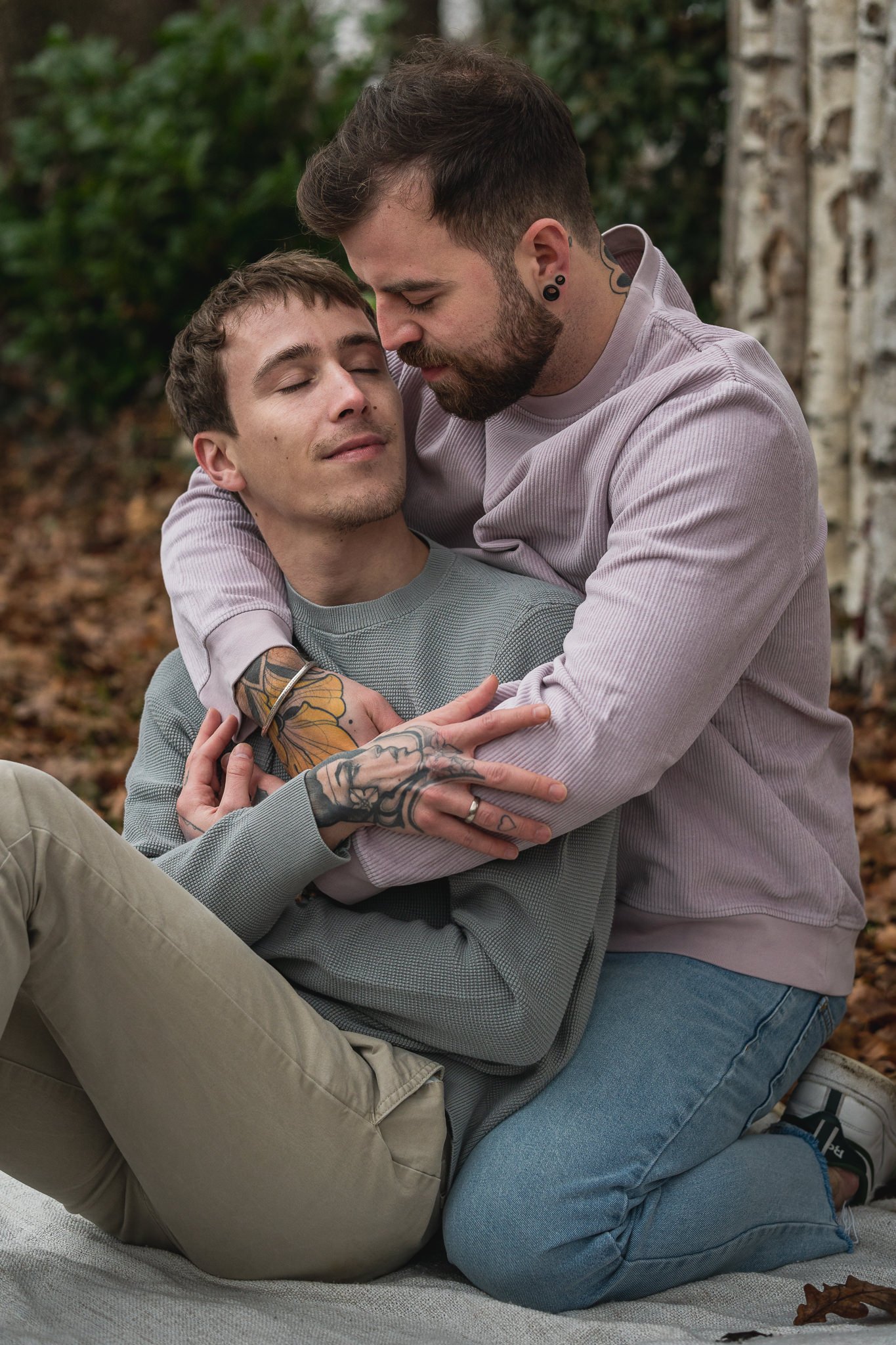Zwei Männer umarmen sich liebevoll im Freien, sitzen auf einem Teppich, umgeben von Herbstblättern und Bäumen.