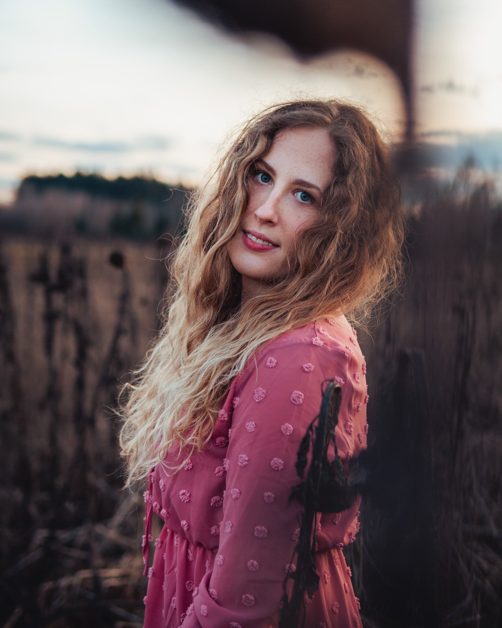 Junge Frau mit langen, lockigen blonden Haaren trägt pinkes Kleid mit Stoffblumen, steht im Freien bei Sonnenuntergang vor dunklem Drahtzaun.