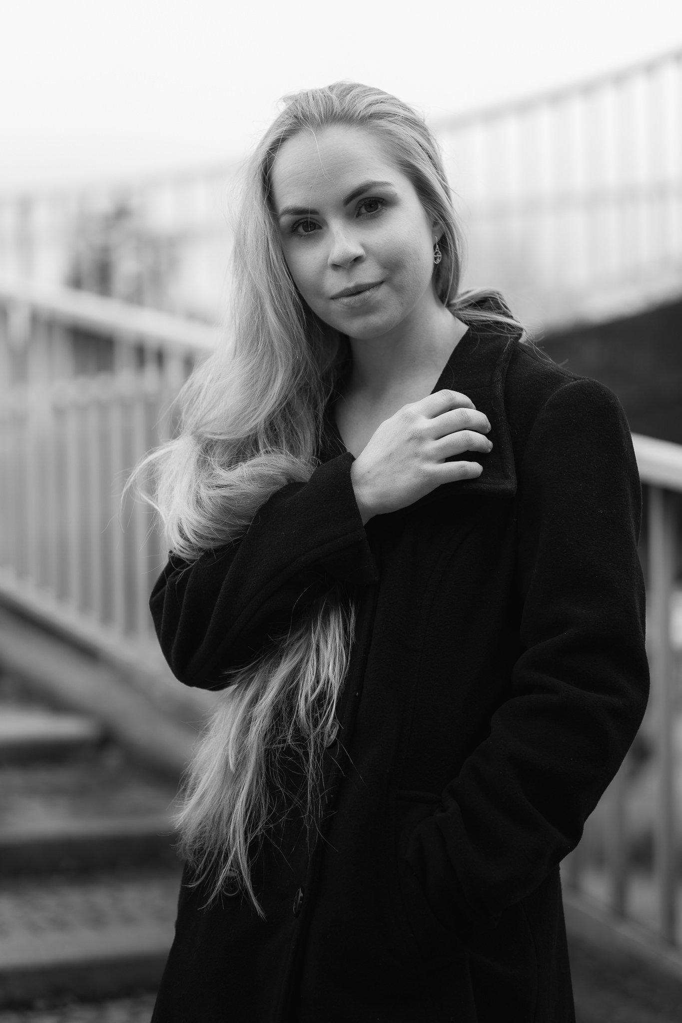 Schwarz-weiß-Foto einer jungen Frau mit langen Haaren, die eine dunkle Jacke trägt und an einem draußen liegenden Bahnhof steht.