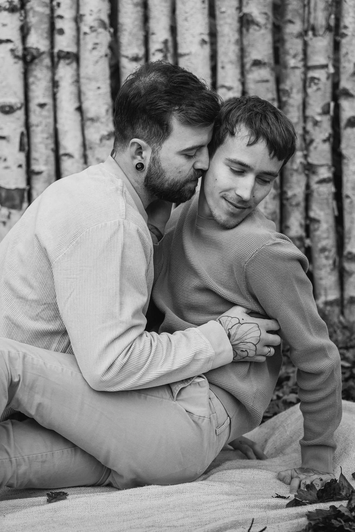 Zwei Männer sitzen eng beieinander auf einem Teppich im Freien vor einem Baumstamm, eines umarmt das andere. Sie scheinen emotional verbunden, einer küsst den anderen sanft an den Kopf.