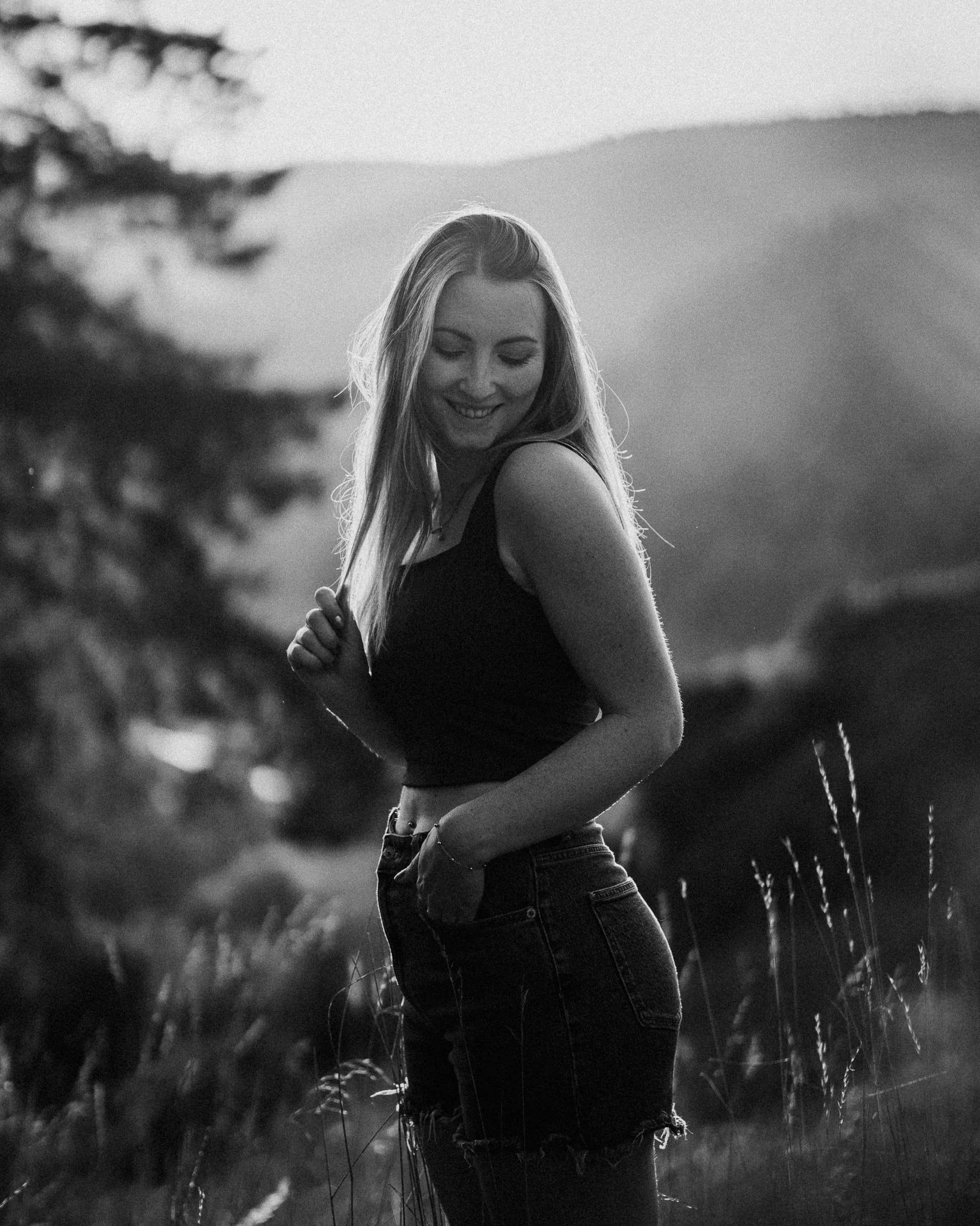 Schwarz-weiß-Foto eines lächelnden Mädchens im Freien, umgeben von hohen Gräsern, bei Sonnenuntergang im Hintergrund.