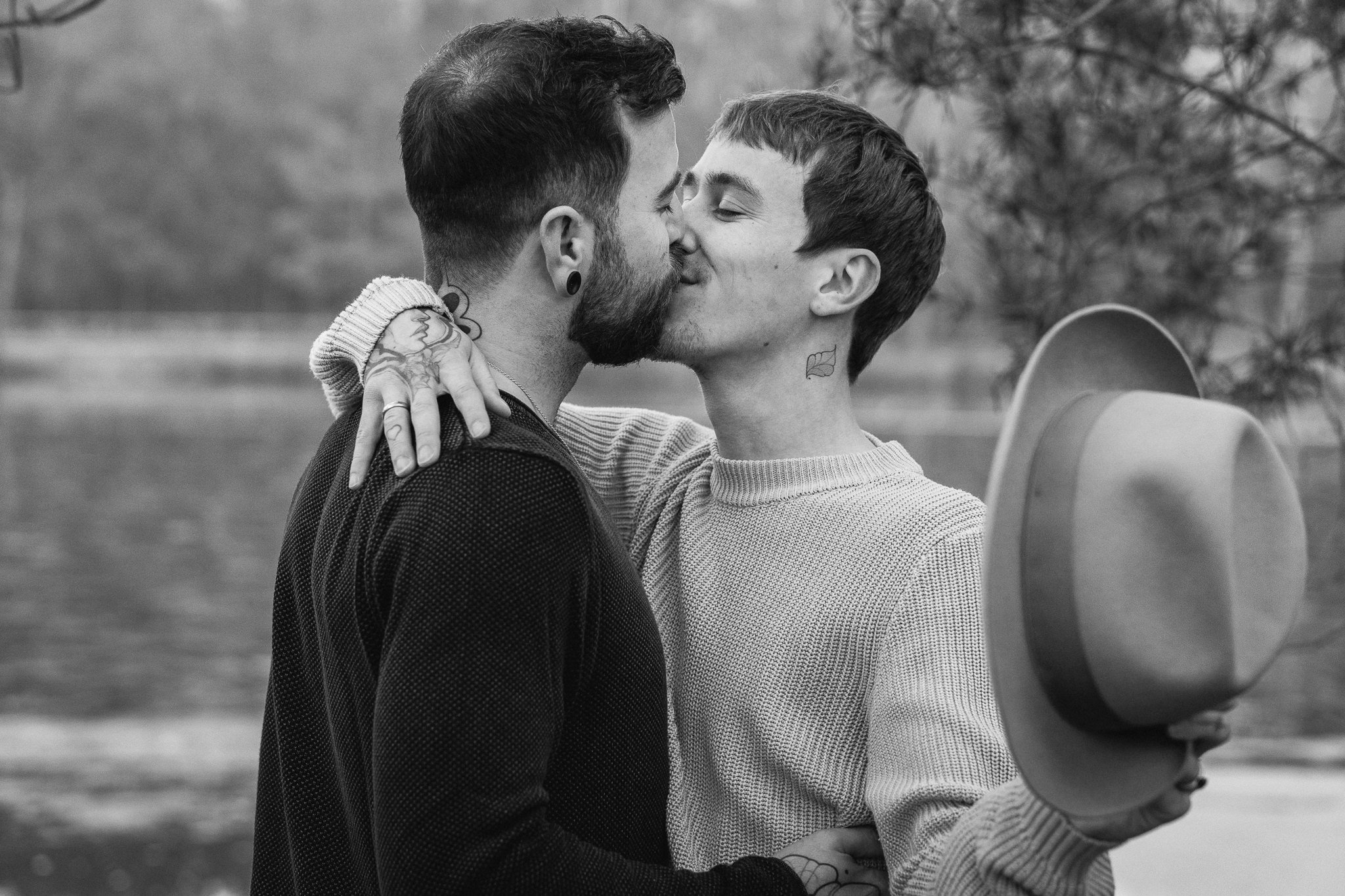 Zwei Männer küssen sich im Park, einer hält einen Hut in der Hand, im Hintergrund Bäume. Schwarz-Weiß-Foto.