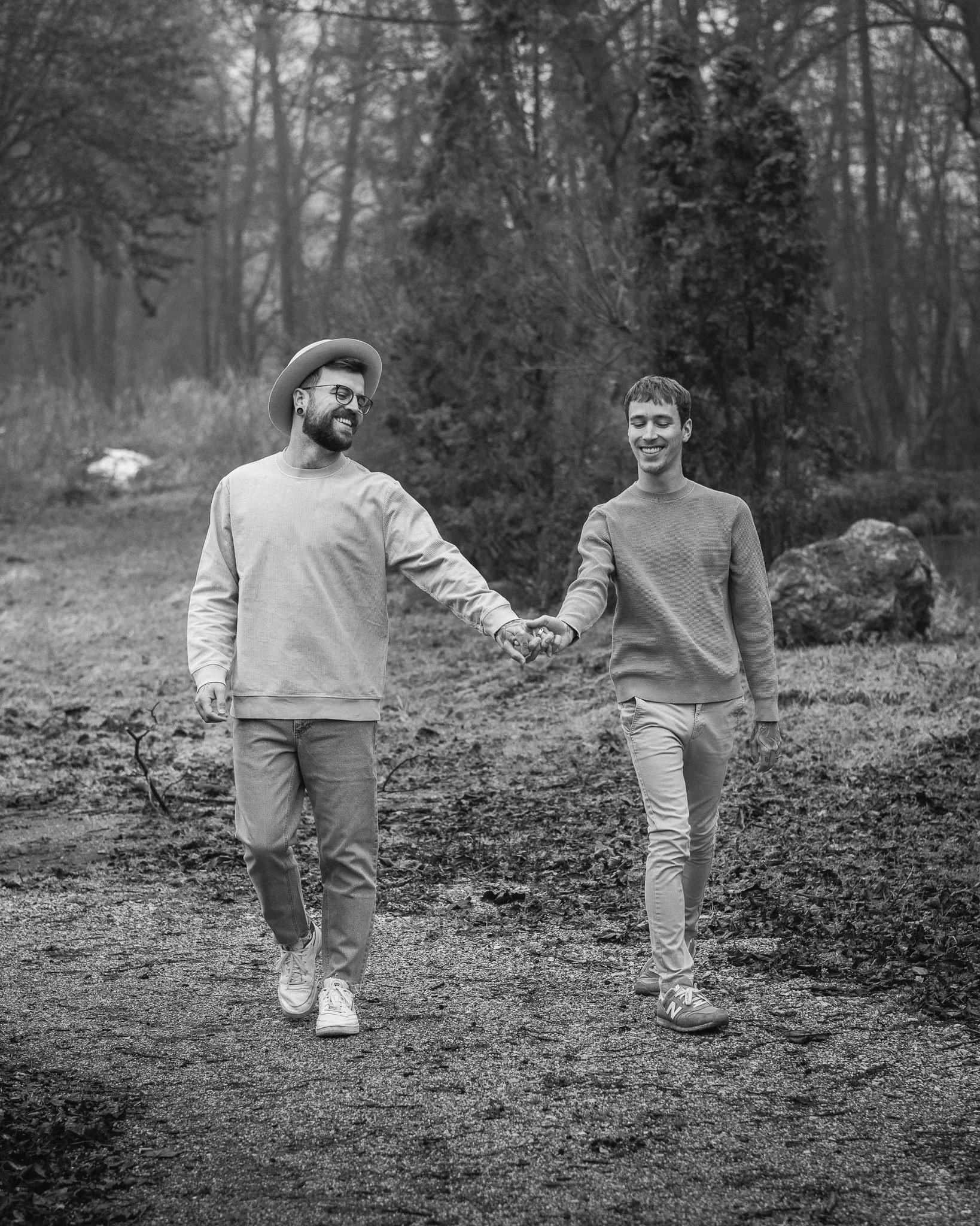 Zwei junge Männer gehen Hand in Hand durch einen Wald, lachen und genießen die Natur.