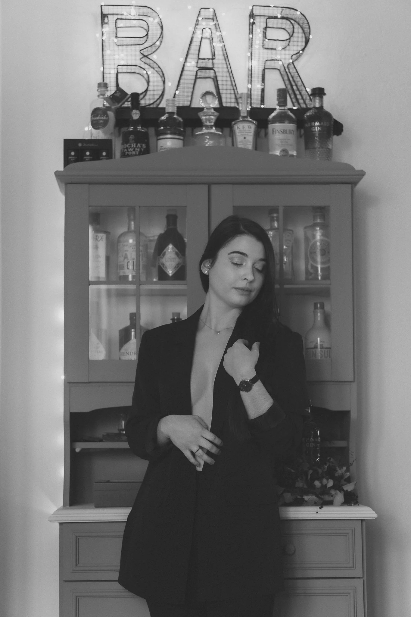 Eine junge Frau in dunklem Blazer vor einer Bar mit beleuchteten Buchstaben, die das Wort 'BAR' bilden, und einer Bar-Garnitur im Hintergrund.