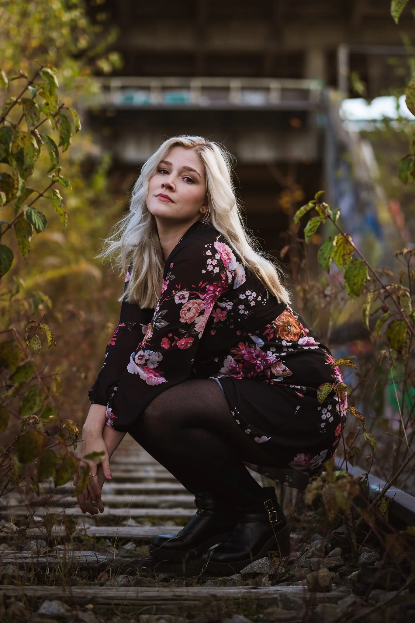 Eine junge Frau mit blonden Haaren, schwarzer Strickjacke und Blümchen-Kleid sitzt auf einem alten Eisenbahn-Schotterweg umgeben von Pflanzen und Büschen, im Hintergrund eine Brücke.
