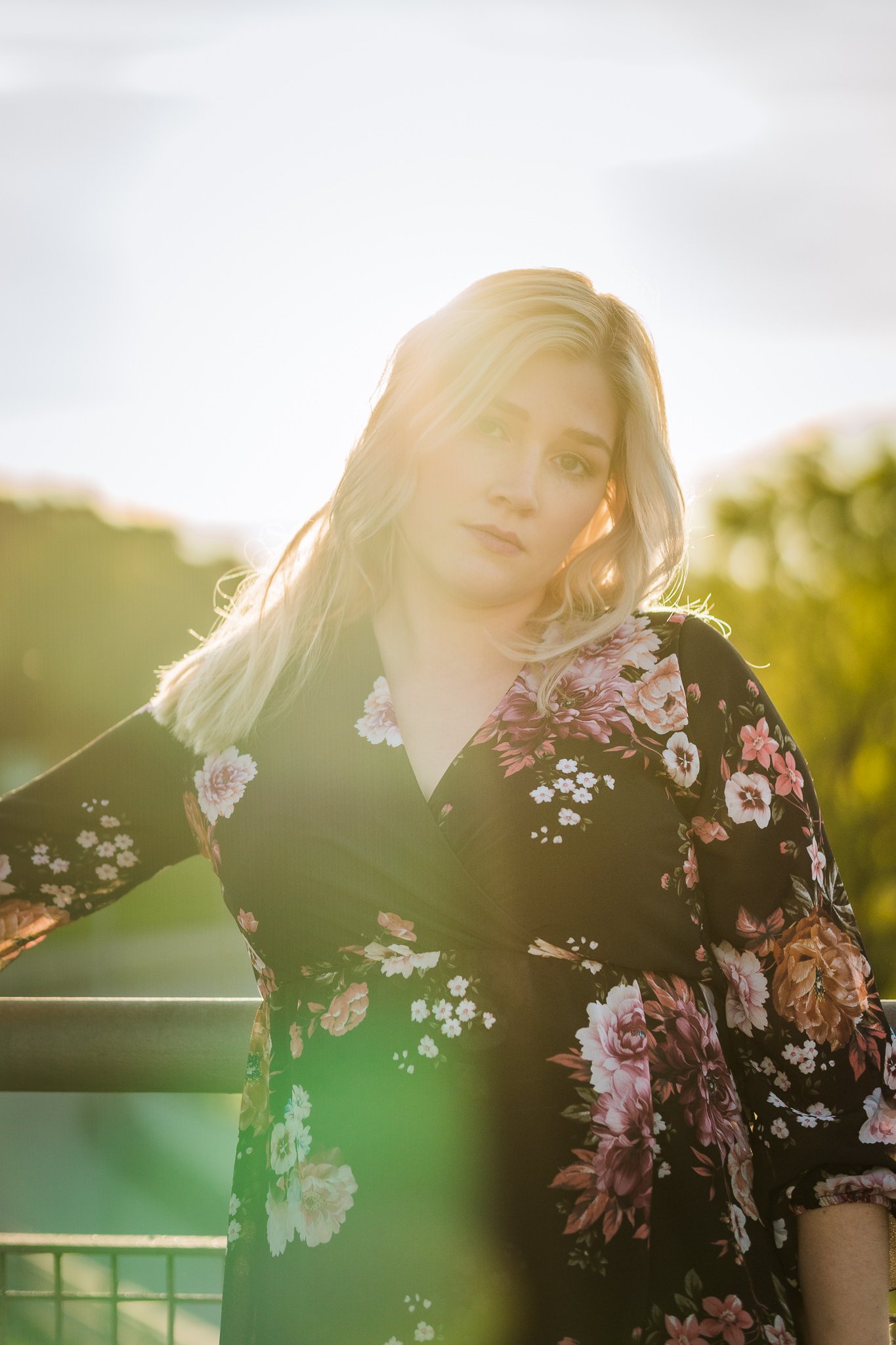 Junge Frau mit blonden Haaren, im Sonnenlicht, trägt ein schwarzes Kleid mit rosa Blumenmuster, steht im Freien.