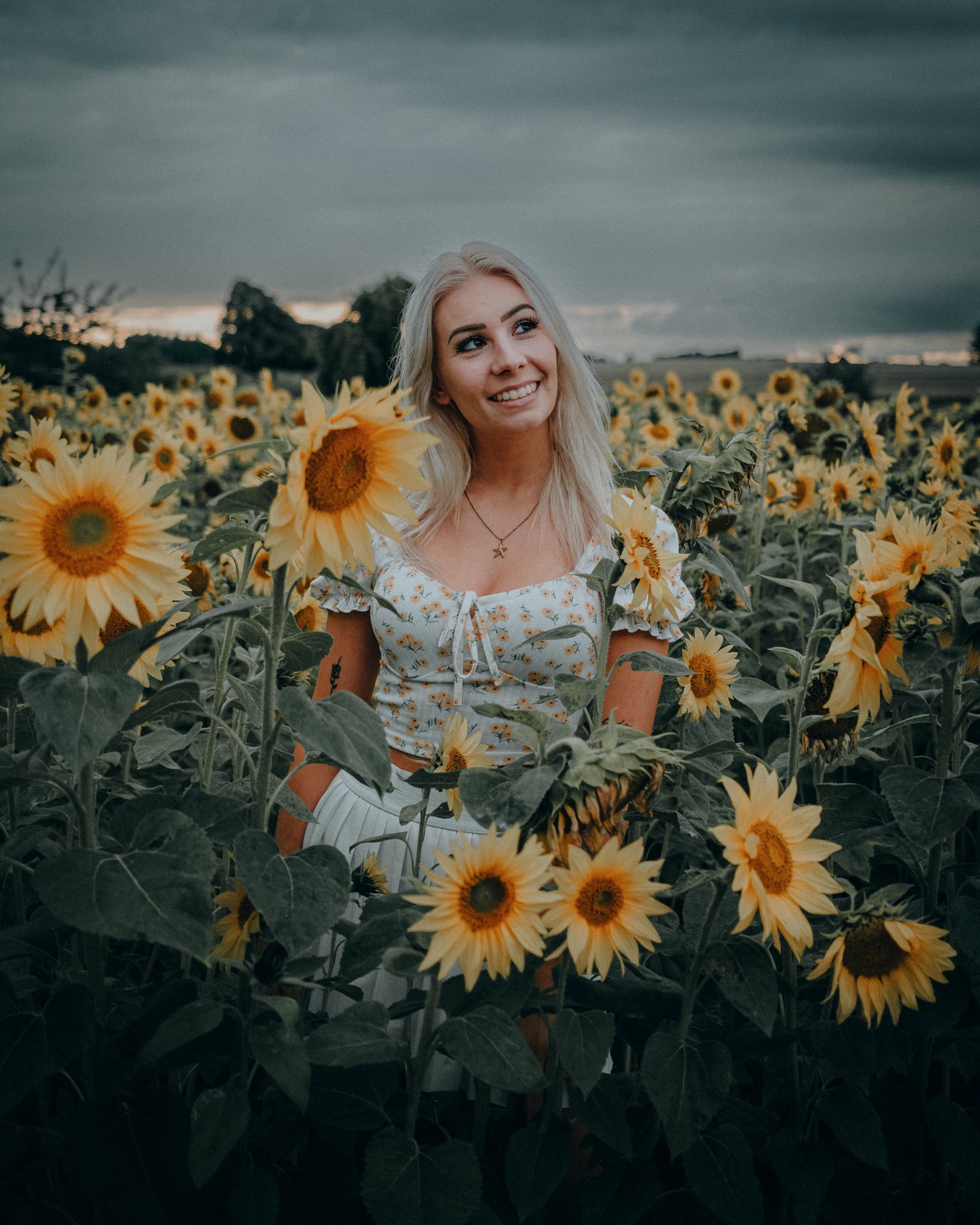 Eine junge blonde Frau lächelt in einem Sonnenblumenfeld bei bewölktem Himmel.