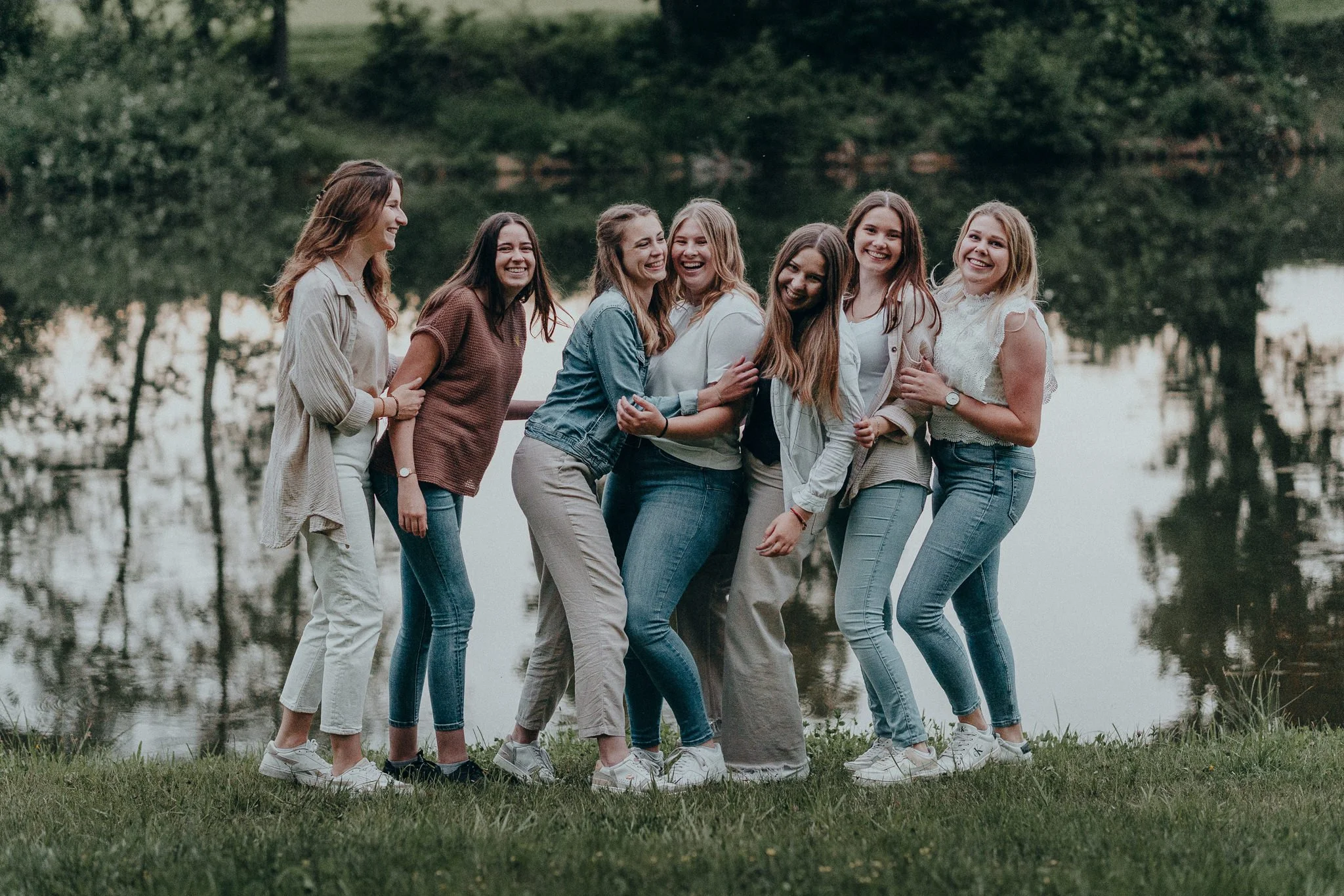 Sieben lachende Frauen stehen am Ufer eines Sees im Grünen, die Landschaft im Hintergrund ist bewaldet und ruhig.