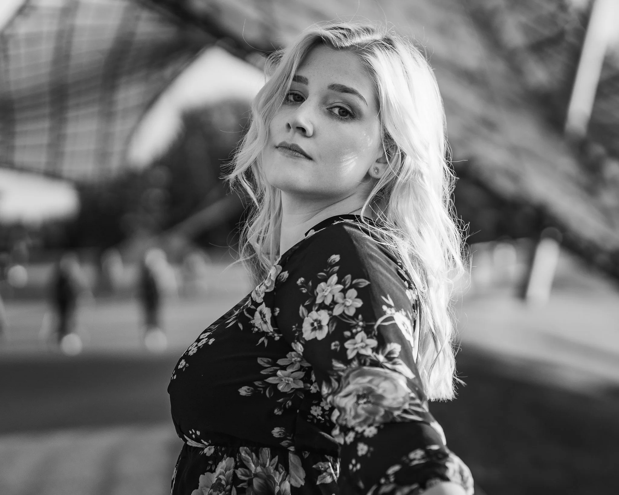 Schwarz-Weiß-Foto einer jungen blonde Frau mit lockigem Haar, die sich leicht zur Seite dreht, vor einem Park mit einem großen Gebäude im Hintergrund.