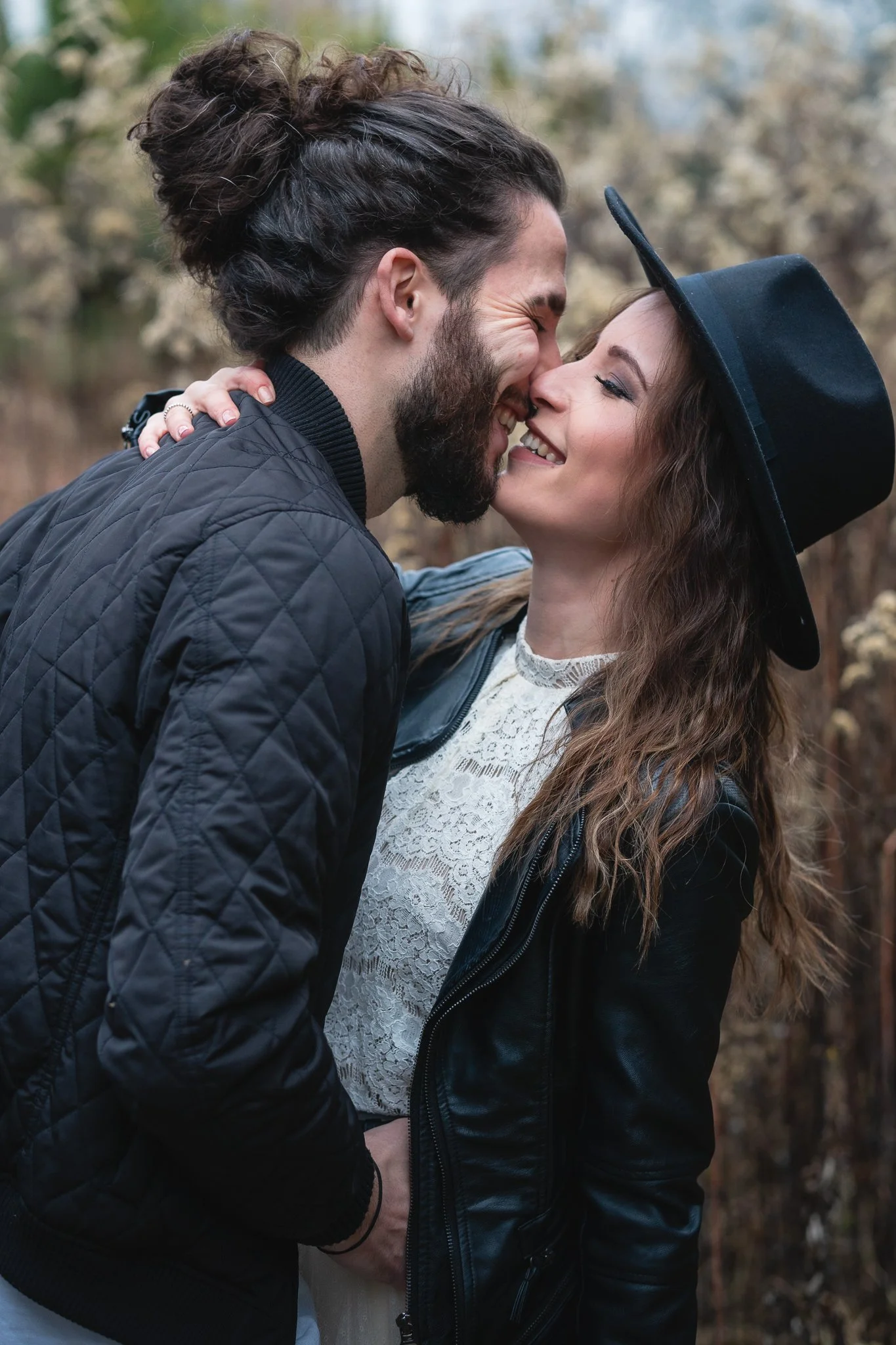 Ein lachendes Paar, das sich liebevoll berührt und sich fast küsst, in einem Außenbereich mit Bäumen im Hintergrund, trägt stilvolle Kleidung inklusive einem schwarzen Lederjacken und einer schwarzen Hut.