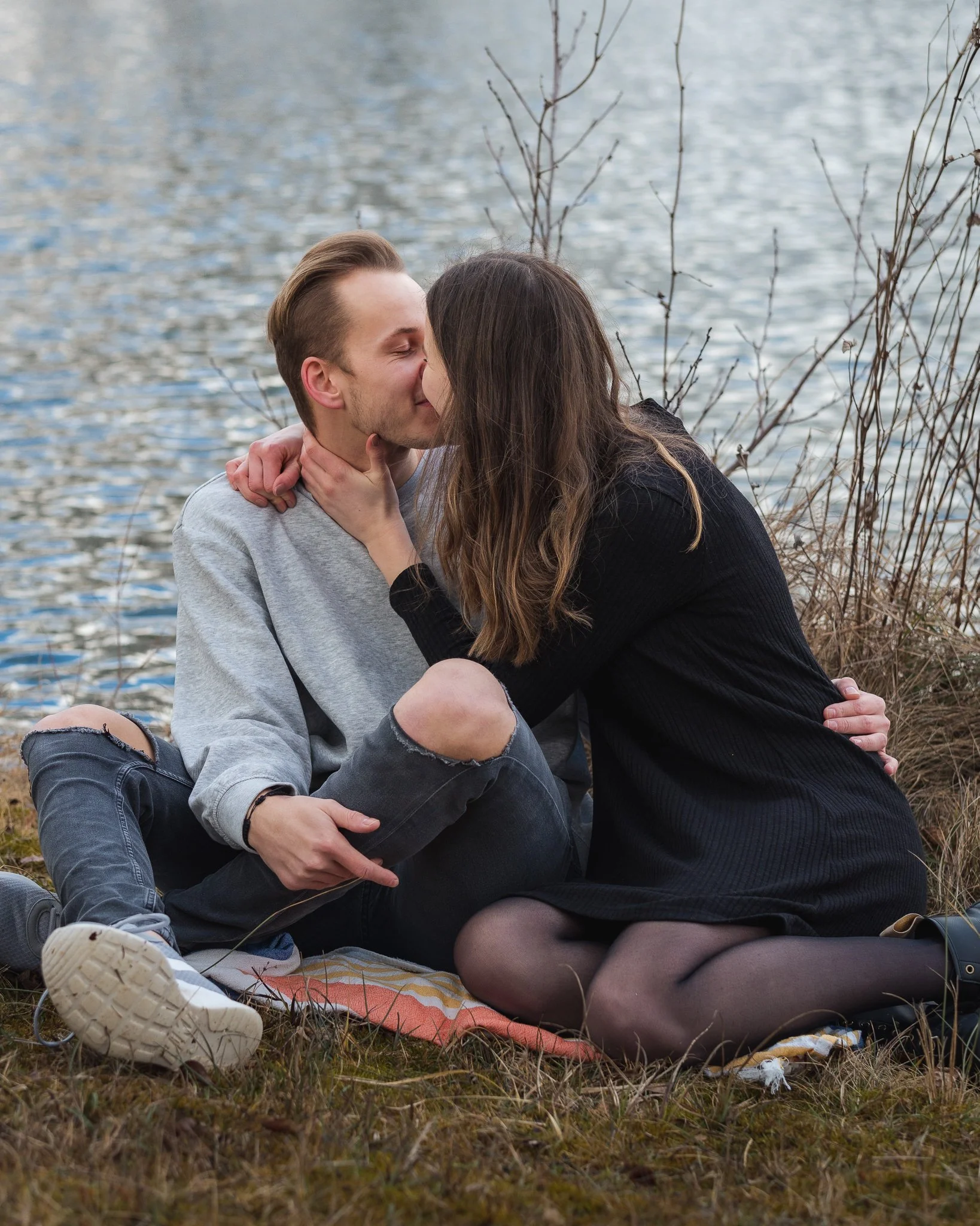 Ein Paar sitzt auf einer Decke am Ufer eines Sees und küsst sich.