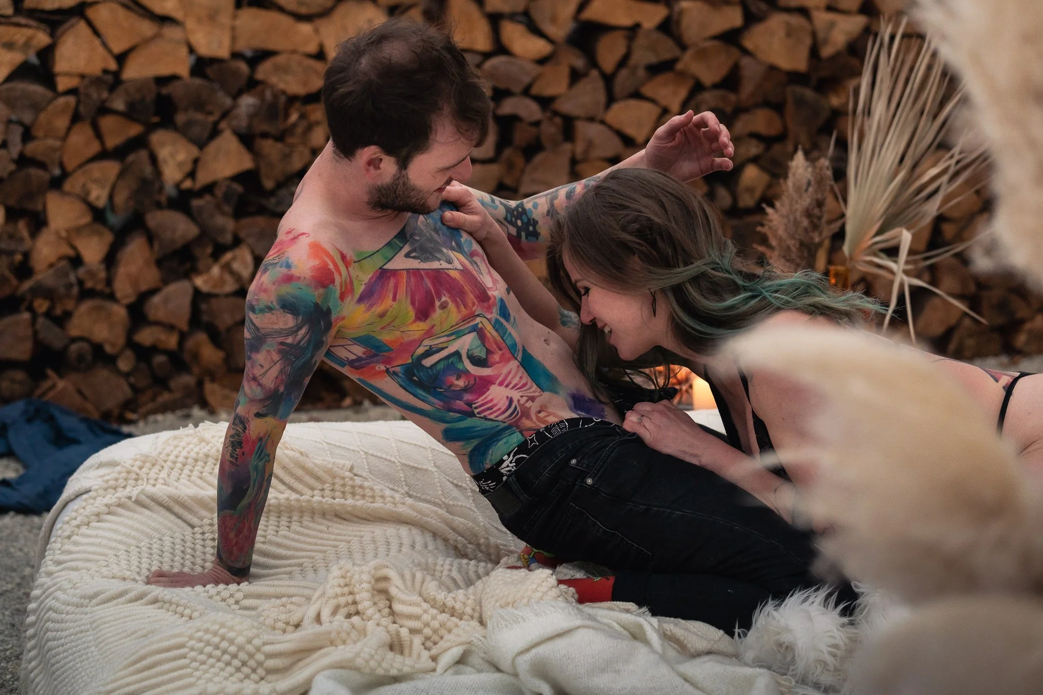 Ein Paar mit Tattoos, das auf einem Bett in einem Raum mit Holzregal im Hintergrund liegt, küsst sich und lacht.