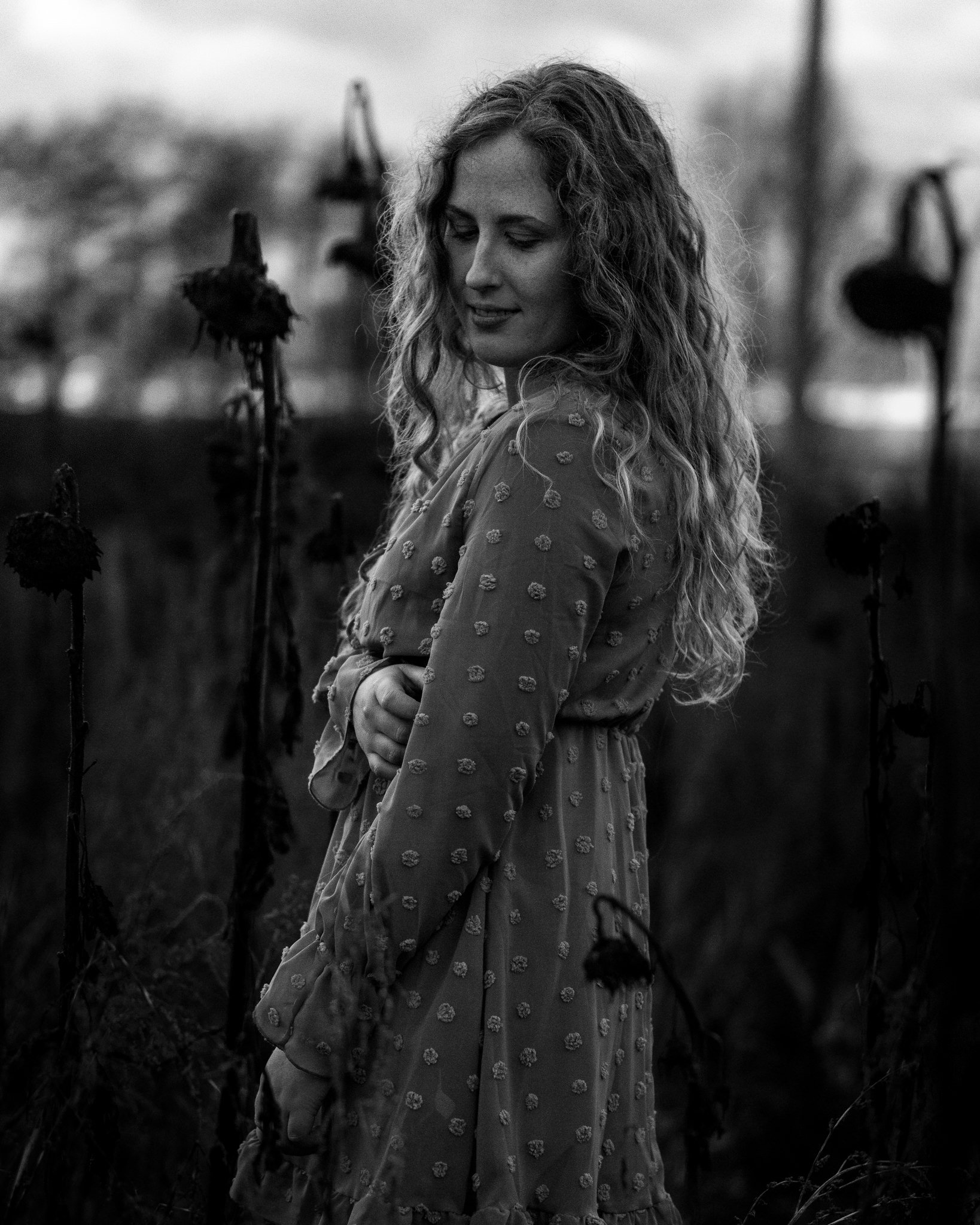 Schwarz-Weiß-Foto einer lächelnden Frau mit lockigem Haar, die eine Seite ihres Kleidungsstammes mit einer Hand hält, im Hintergrund vertrocknete Blumen.