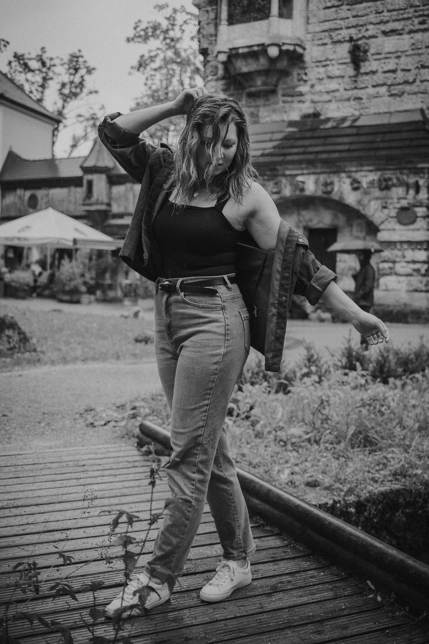 Schwarz-Weiß-Foto einer jungen Frau, die auf einem Holzsteg steht, mit einem alten Gebäude im Hintergrund. Sie trägt Jeans, Sneakers, ein tank top und eine Jacke. Sie schaut nach unten, mit einer Hand im Haar.