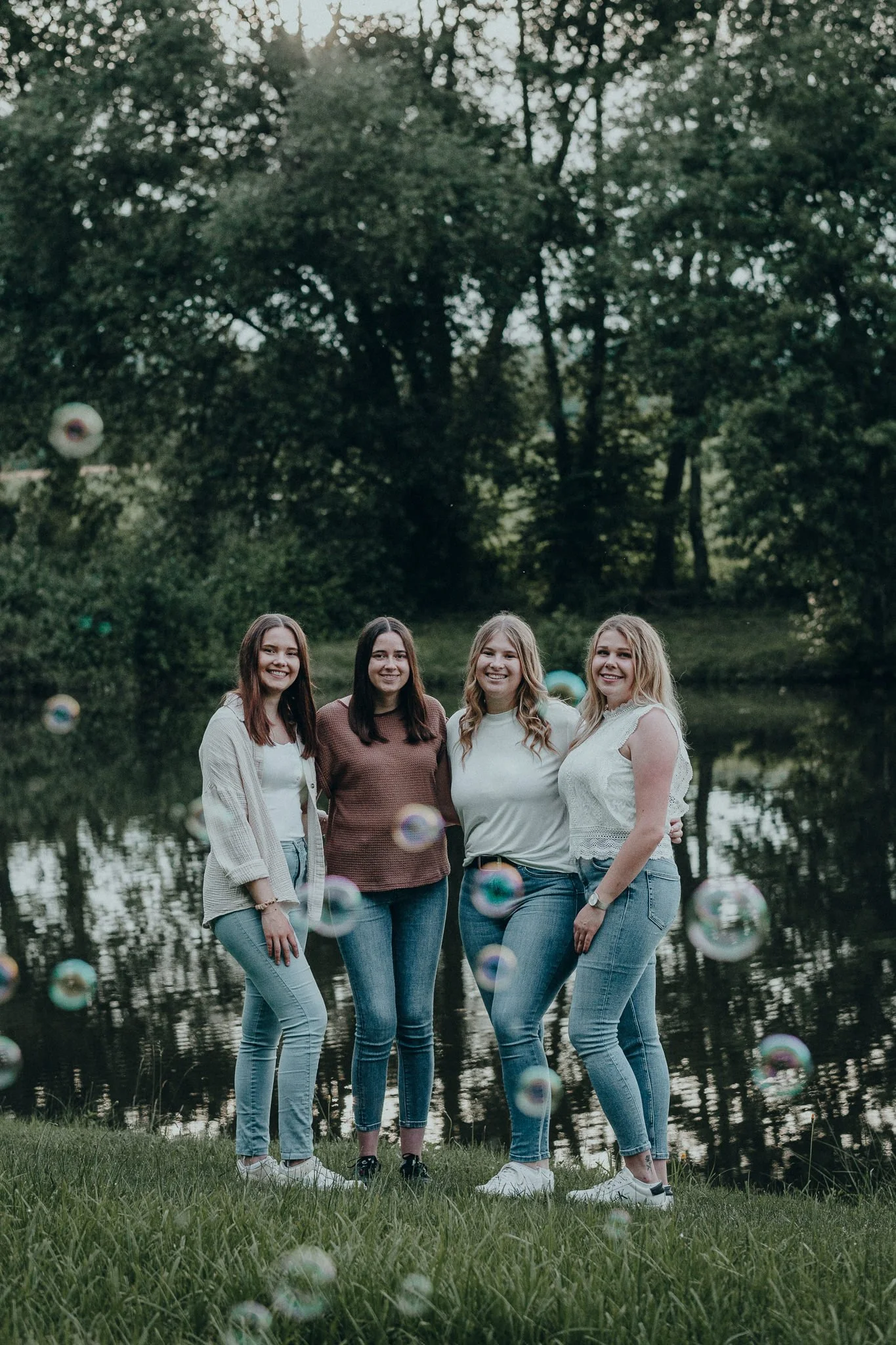 Vier Frauen stehen am Seeufer, umgeben von Bäumen, mit Seifenblasen vor ihnen, das Bild ist bei Sonnenuntergang aufgenommen.
