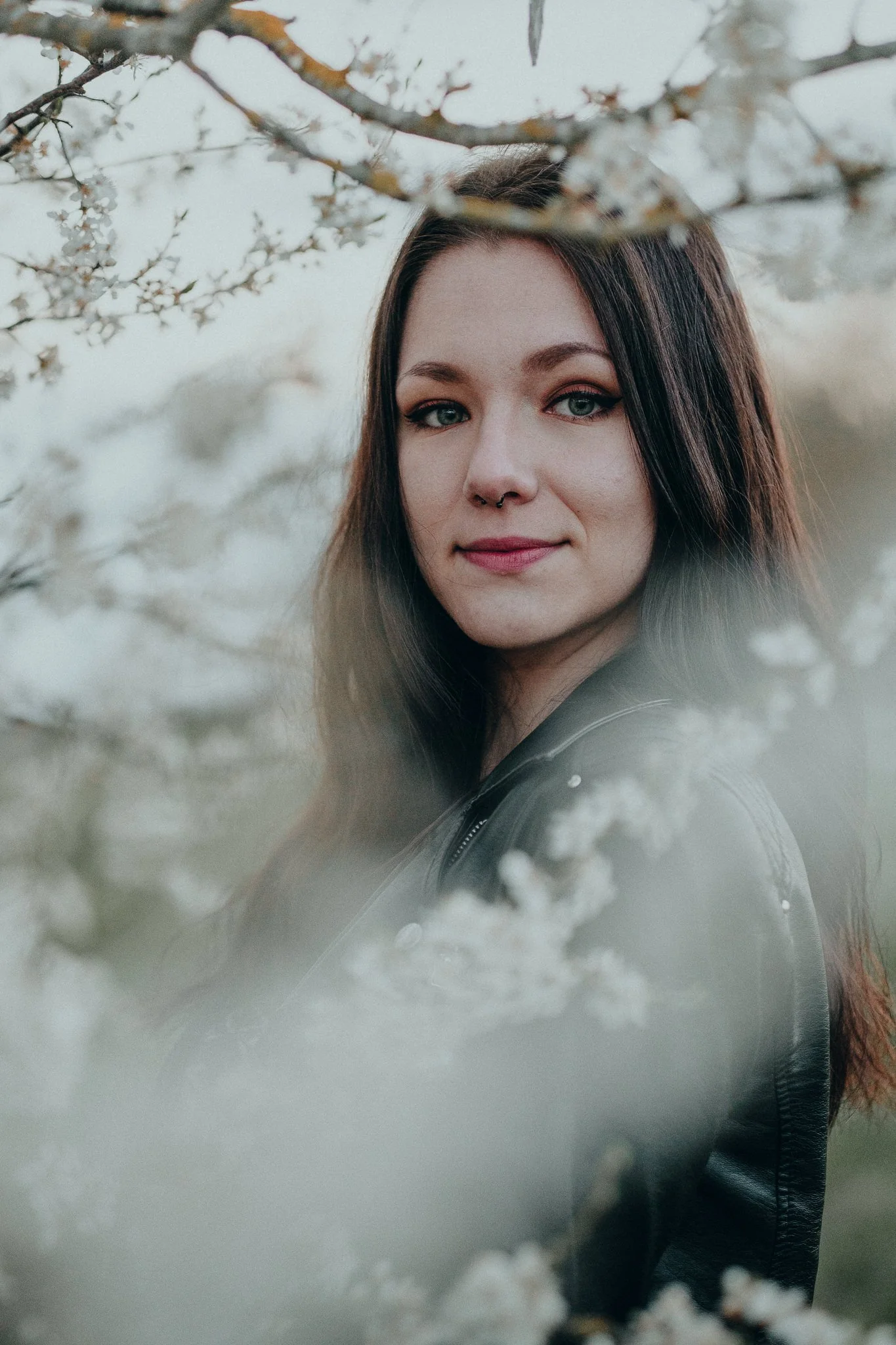 Porträt einer jungen Frau mit dunklem Haar, die zwischen blühenden Zweigen steht. Der Hintergrund ist unscharf, Betrachter sieht nur die Frau mit leichten Makeup und Lederjacke.