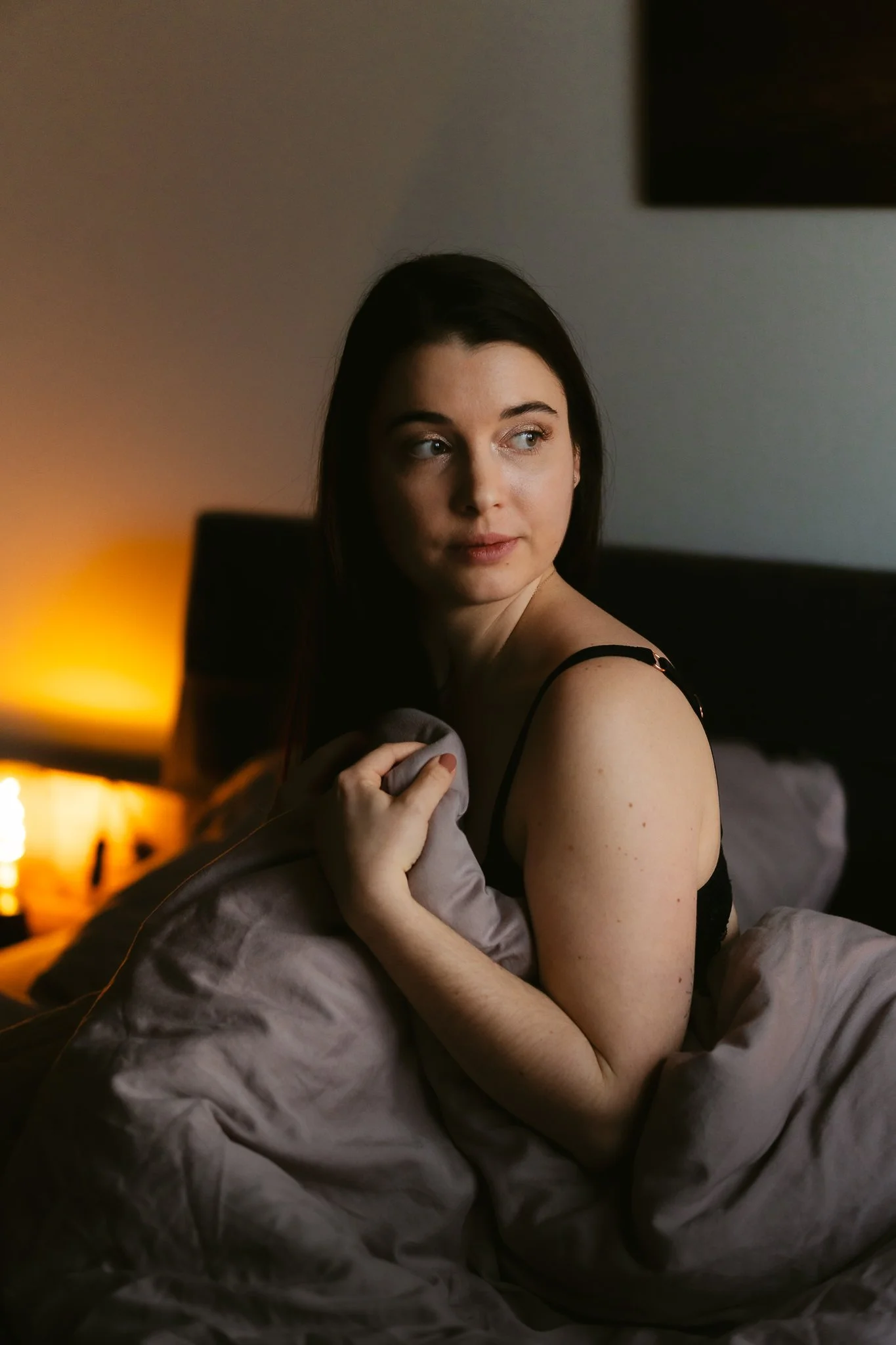 Junge Frau sitzt im Bett, trägt ein schwarzes Nachthemd, hält eine Decke und schaut nach links, im Hintergrund ein warmes Licht.